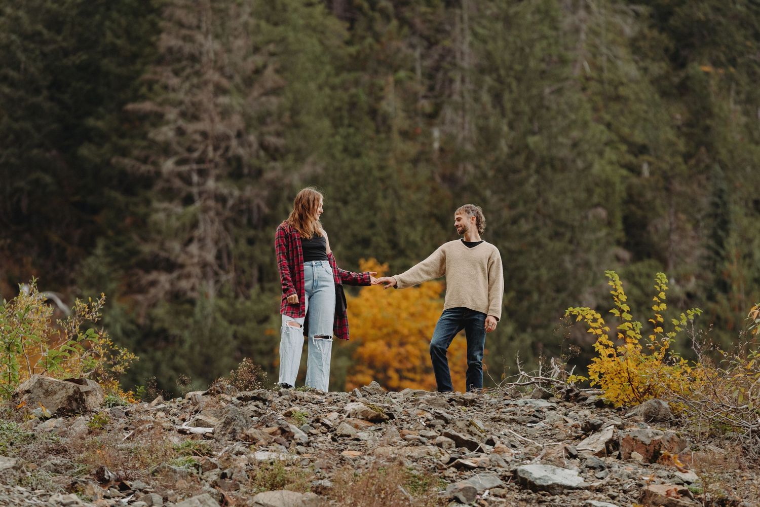 Couple holding hands on rocky terrain surrounded by autumn trees in a mountain forest setting.