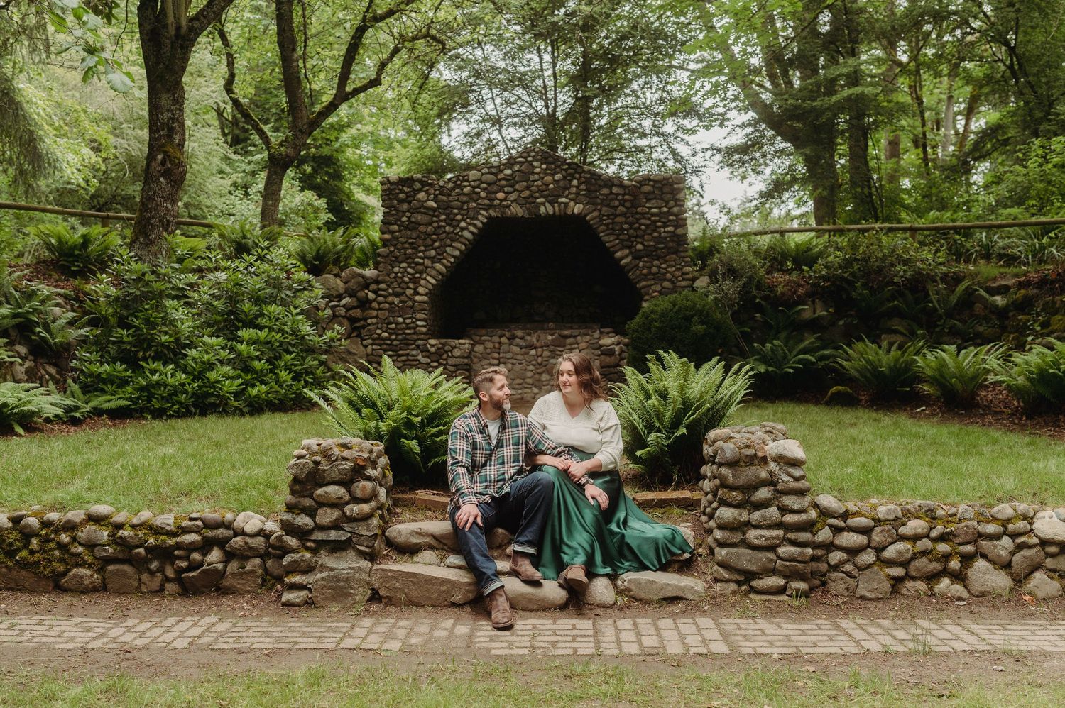 Couple sitting on stone wall in lush garden with rustic grotto backdrop for outdoor engagement photos.
