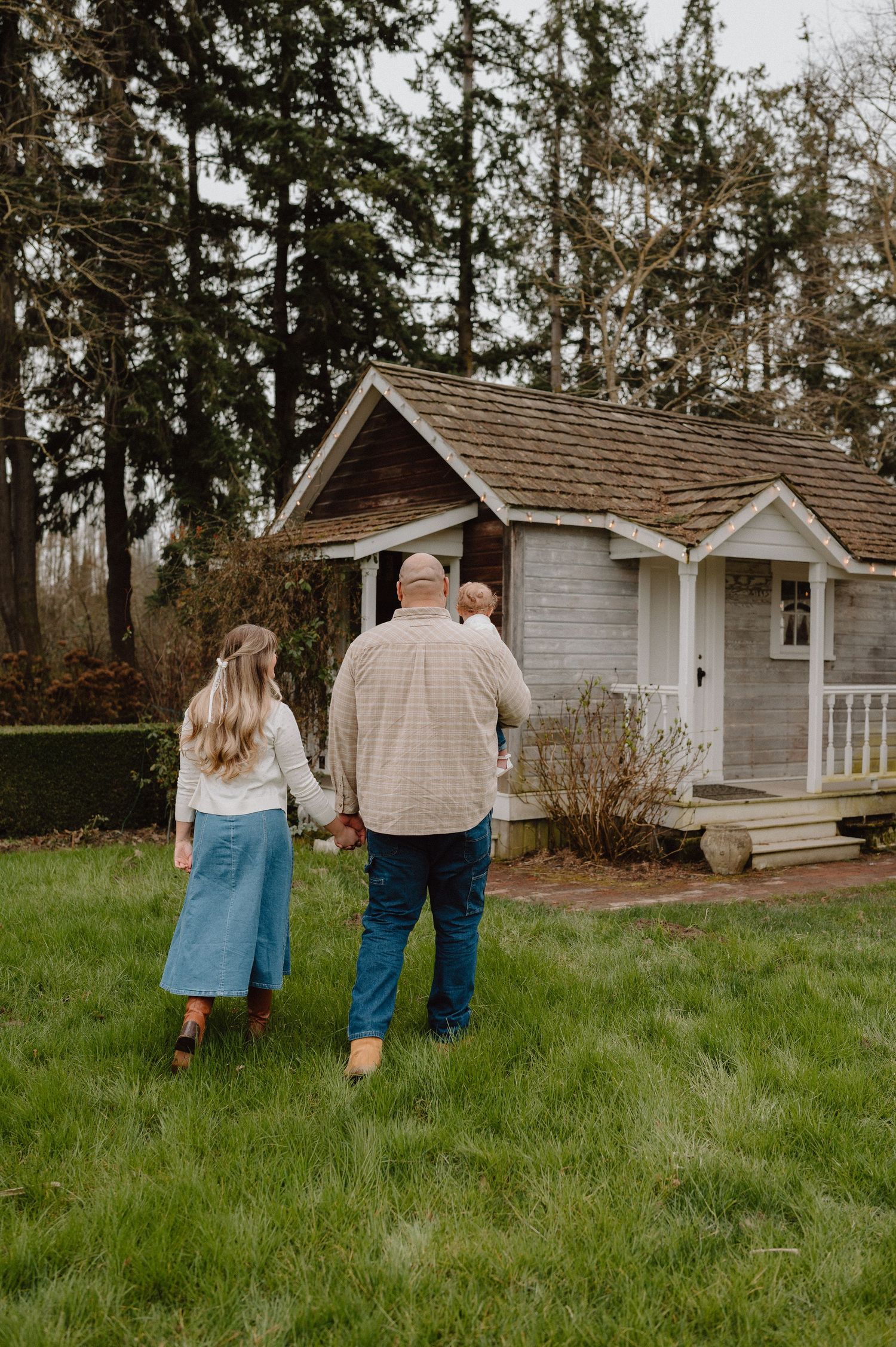 A family of three walks toward a charming rustic cottage surrounded by tall trees and green grass.