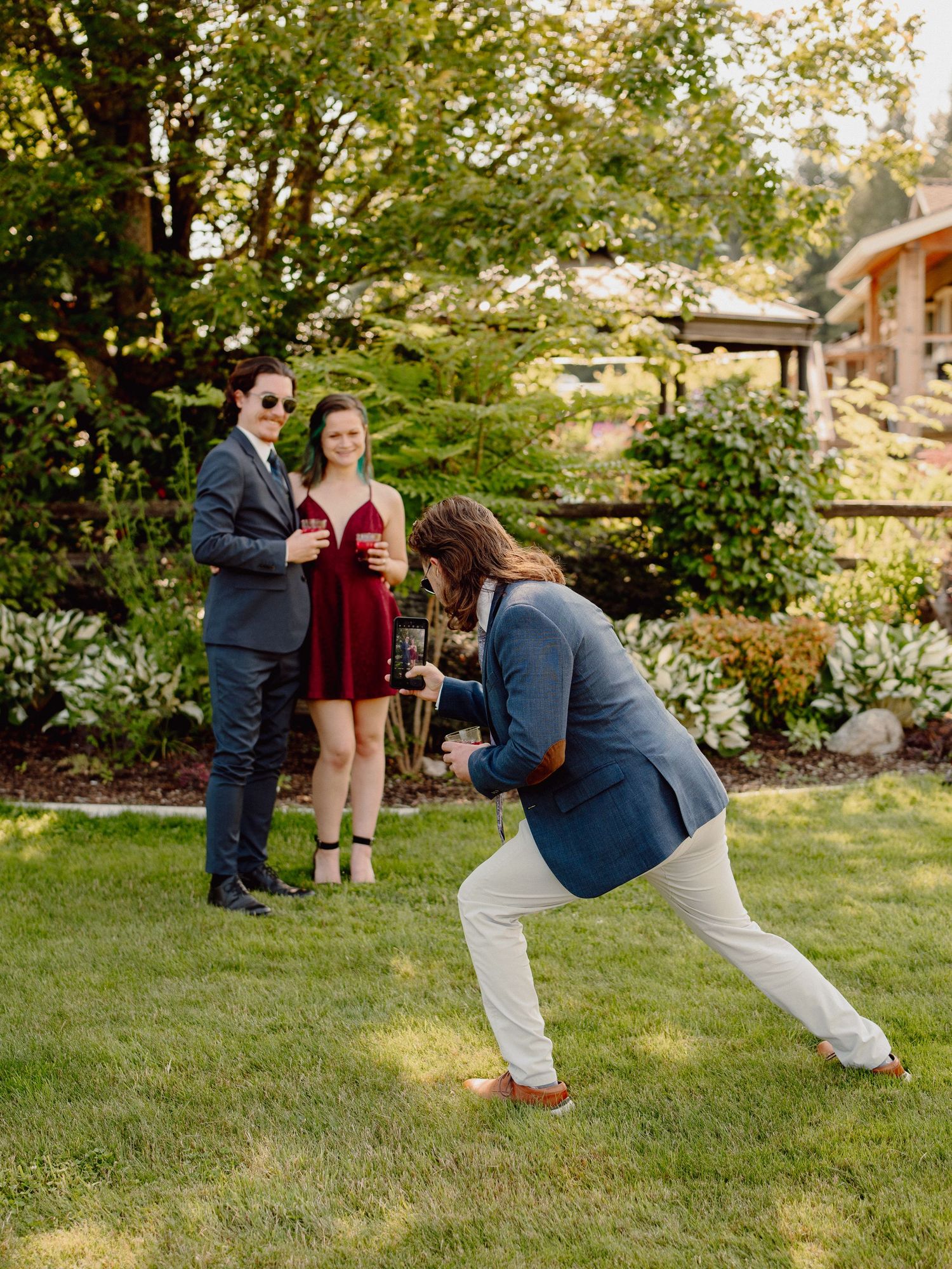 A photographer crouches low to capture a couple in formal attire standing on a manicured lawn.