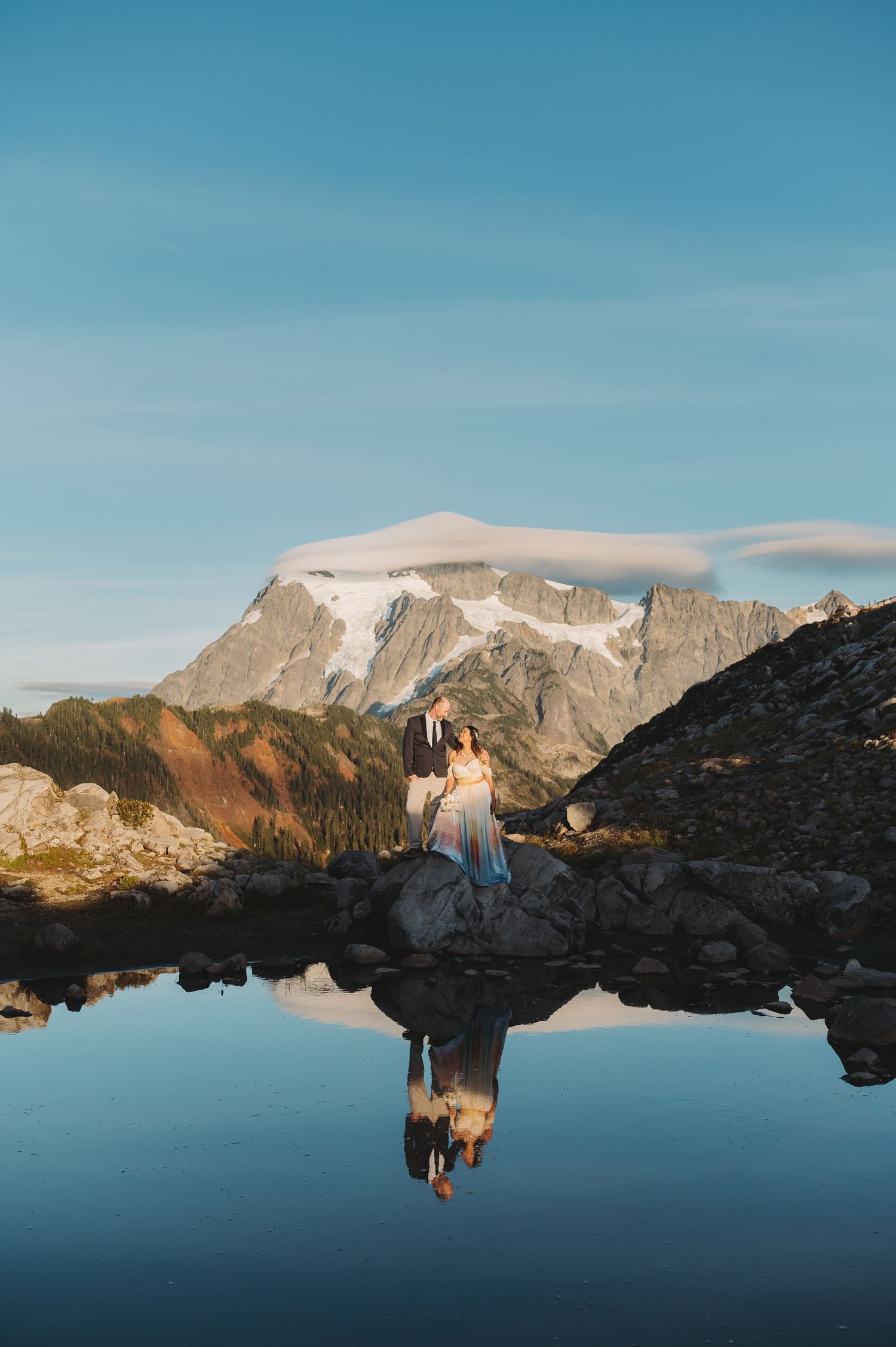 A figure in a white dress stands near a mountain lake, creating a perfect reflection in the still water.