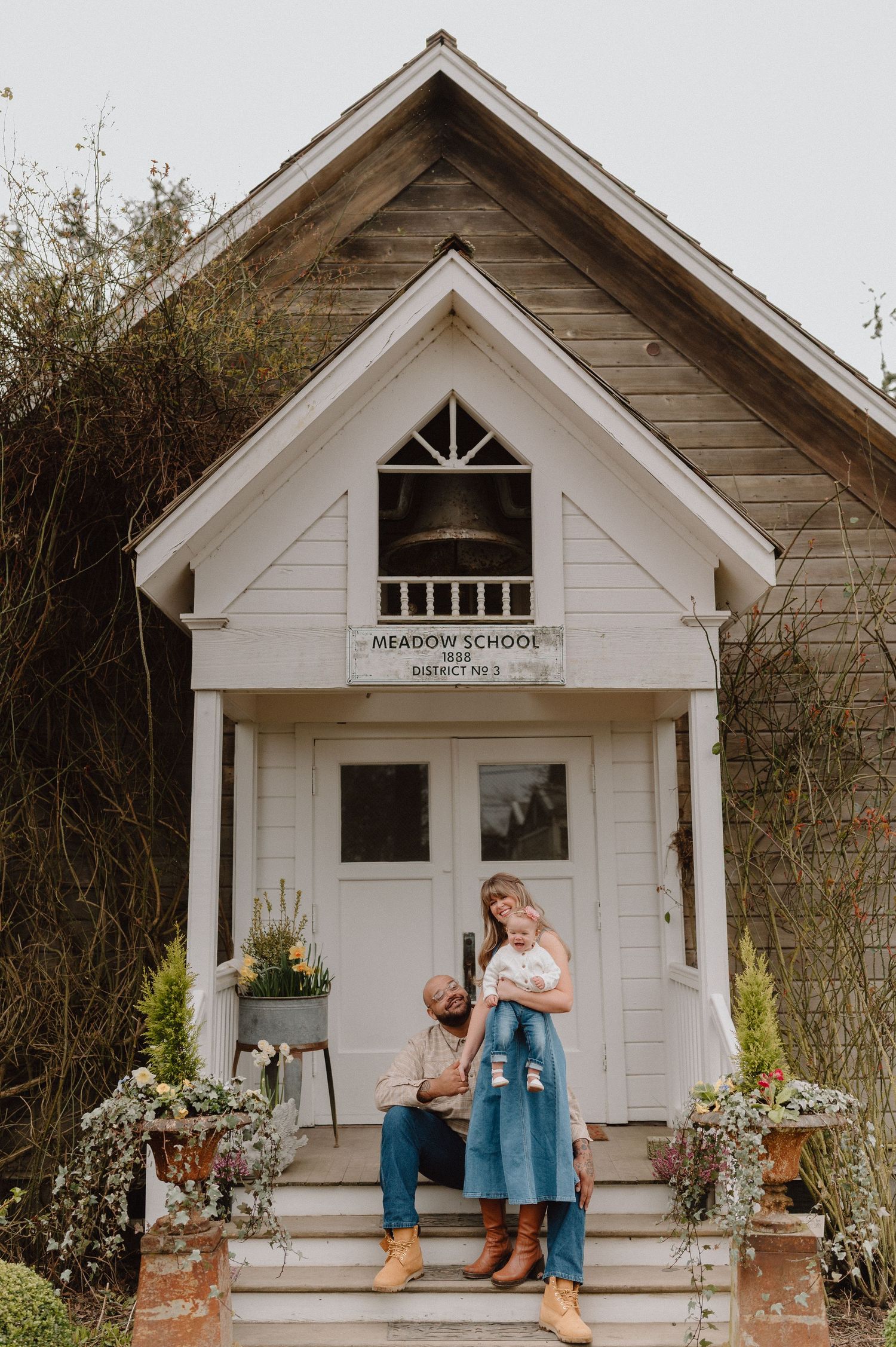 A couple with a baby poses on the steps of a charming white Meadow School District building surrounded by wildflowers.