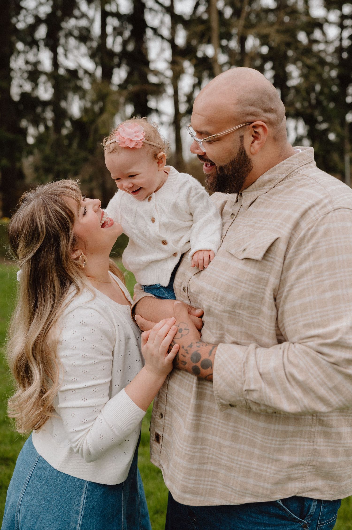A happy young family with a tattooed dad, smiling mom, and baby girl in a pink bow posing outdoors among green trees.