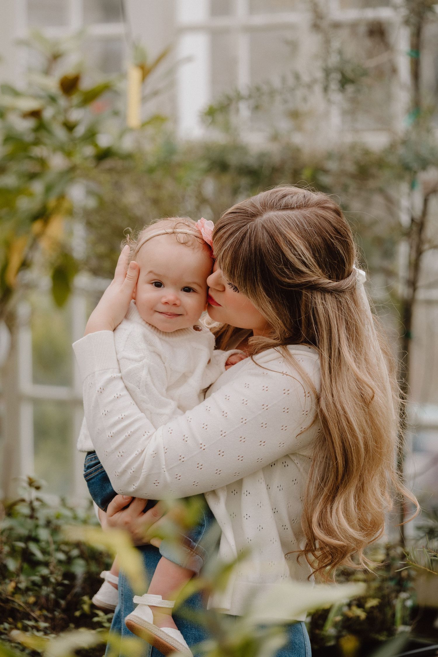 A mother with long blonde hair kisses her smiling baby girl outdoors, both wearing white tops.