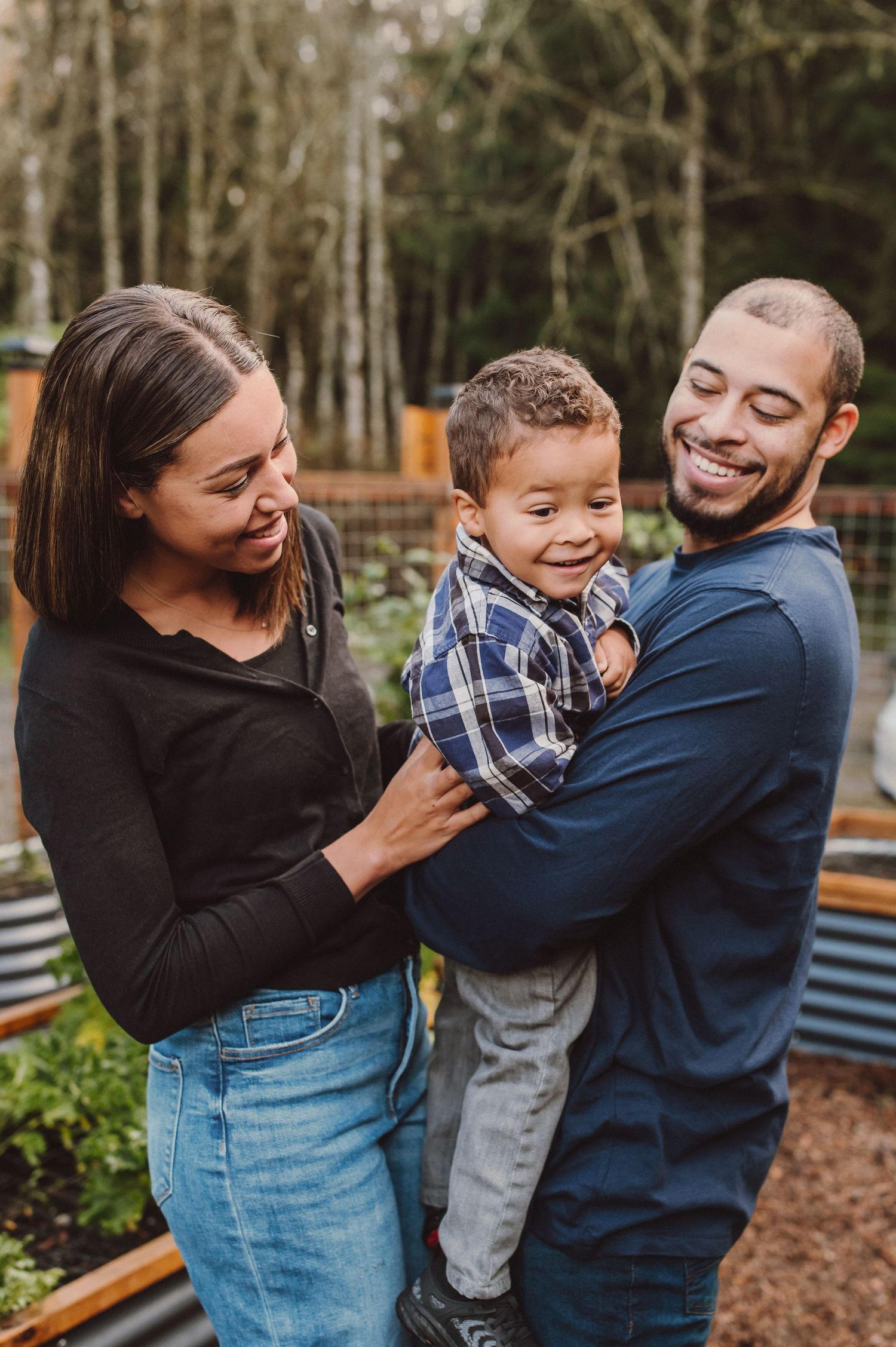 A loving family shares joyful laughter together while spending time outdoors near trees and garden beds.