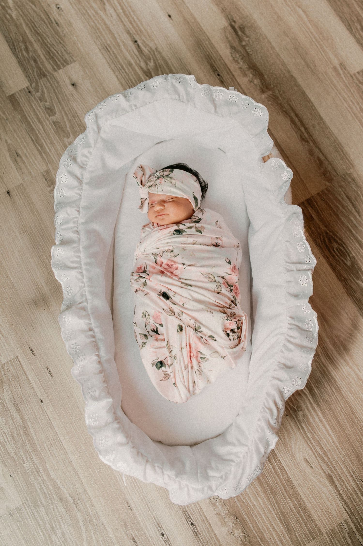 A sleeping newborn swaddled in floral print fabric lies in a white ruffled bassinet on hardwood flooring.