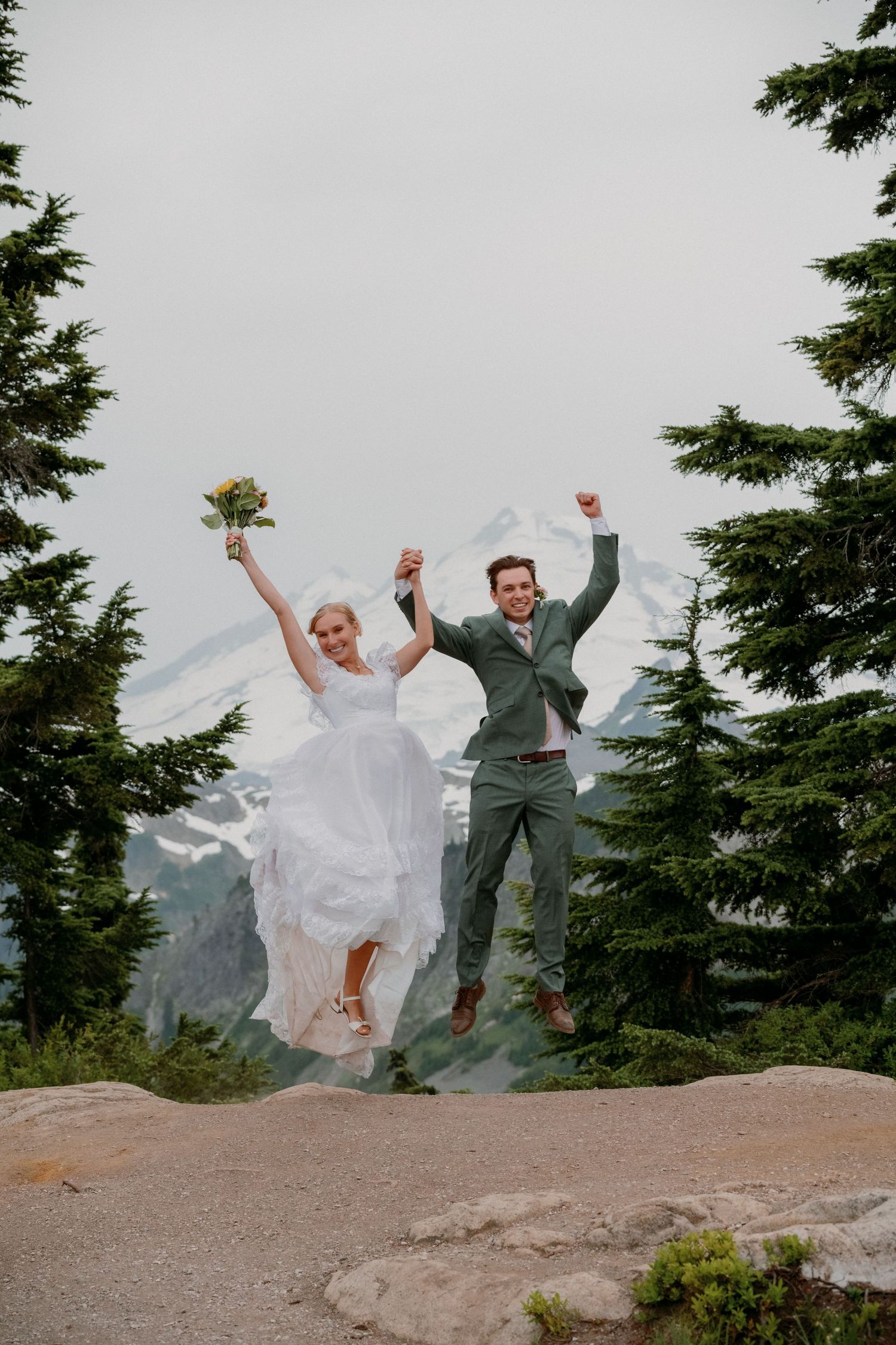A newlywed couple jumps joyfully on a mountain peak surrounded by evergreen trees and snowy mountain views.