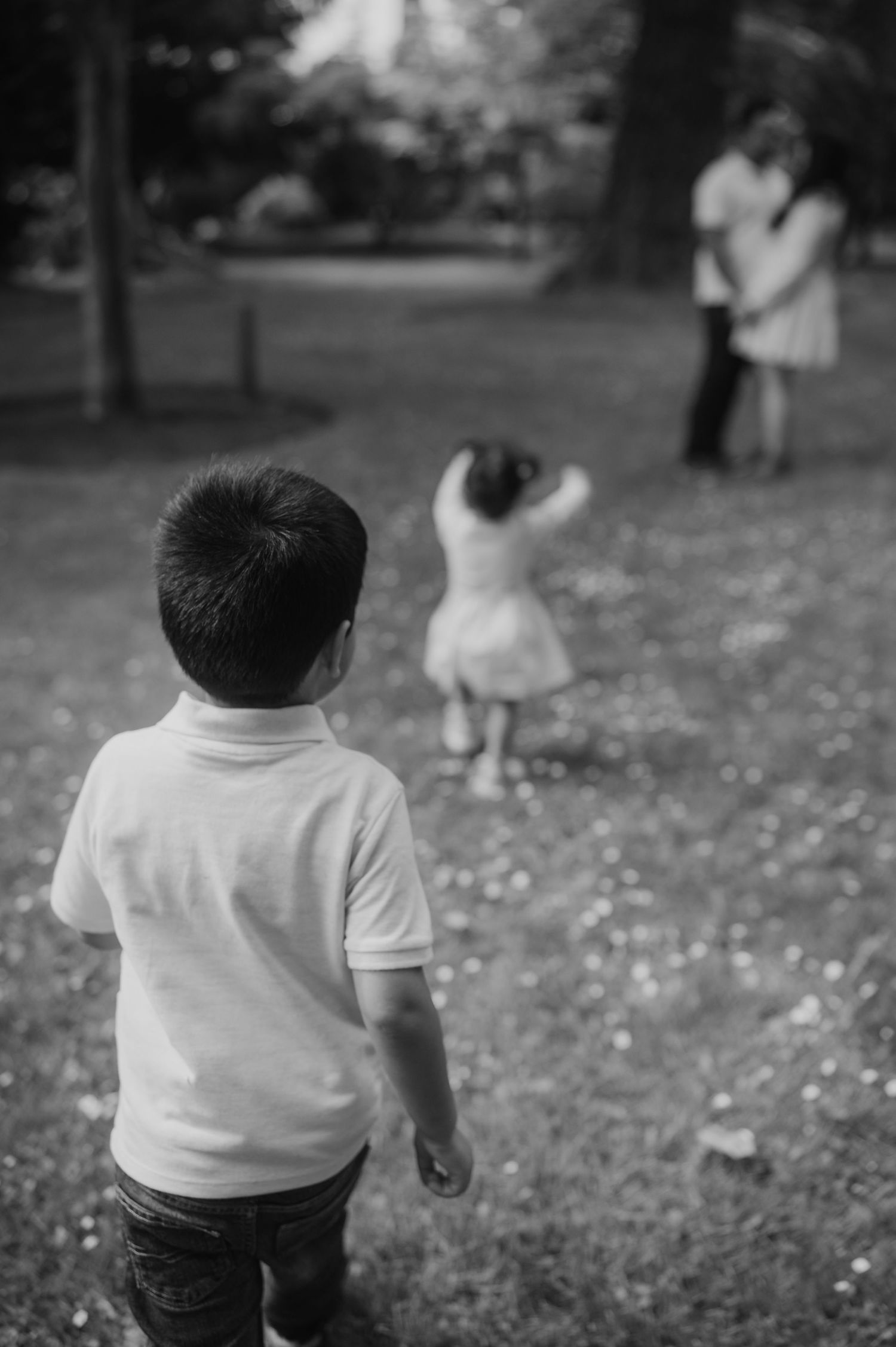 Black and white photo of two children walking toward their parents embracing in a lush green park.