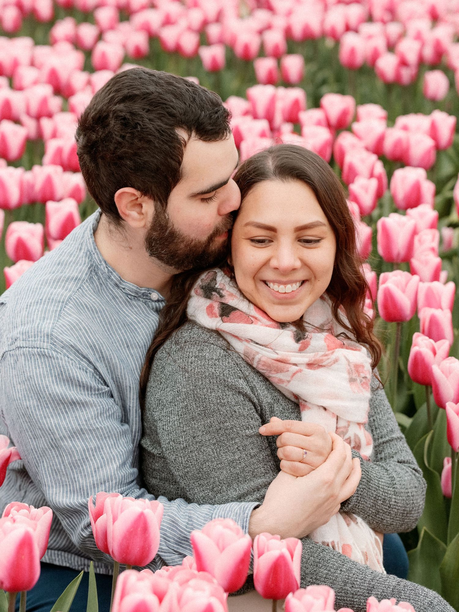 A couple embraces lovingly among pink tulip fields in spring, wearing cozy sweaters during a romantic photo session.