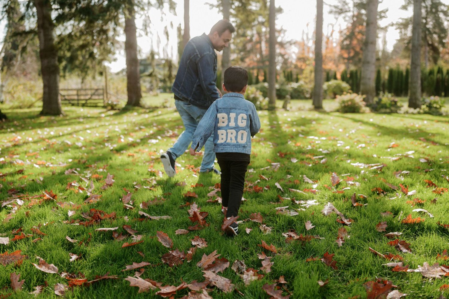 A couple walks together on grass covered with fallen autumn leaves in a park setting.