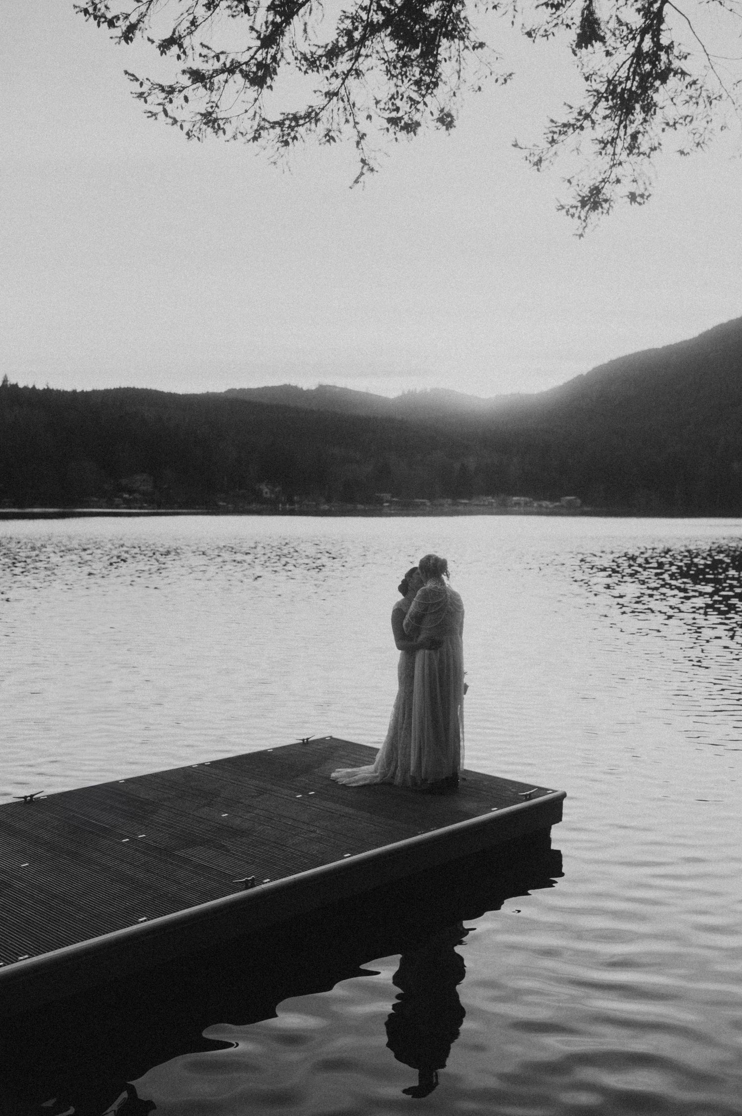 Moody black and white photo of couple embracing on dock overlooking misty mountain lake.