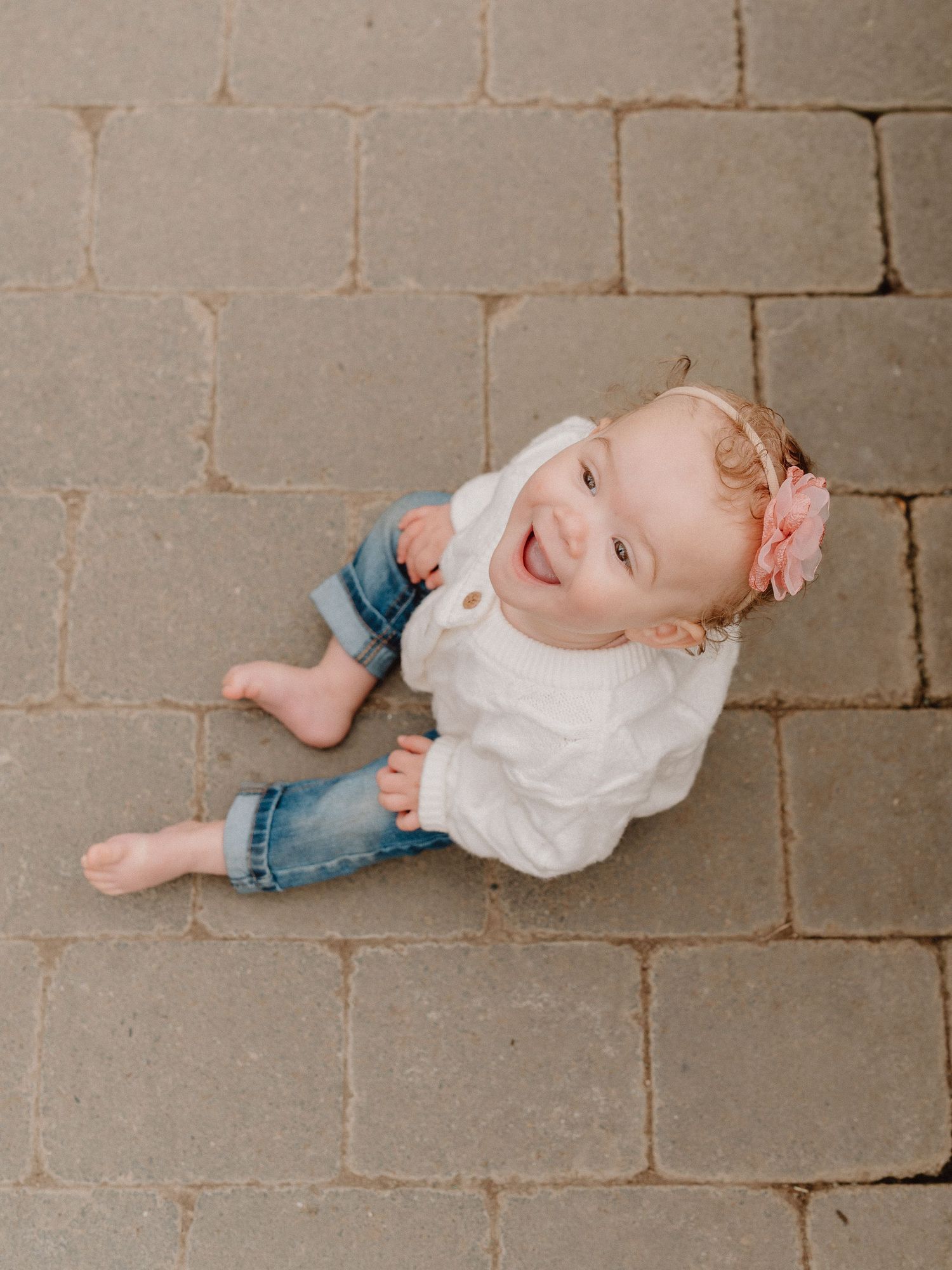 Smiling baby girl in white top and jeans with pink flower headband sitting on brick pavers looking up.