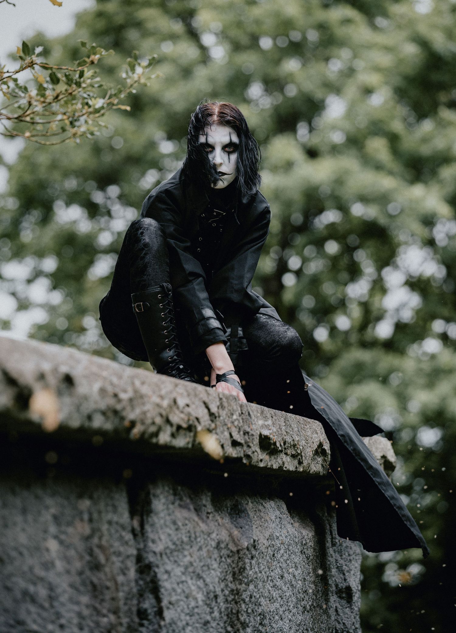 A dark figure in gothic attire sits perched on a stone wall against a blurred green foliage background.