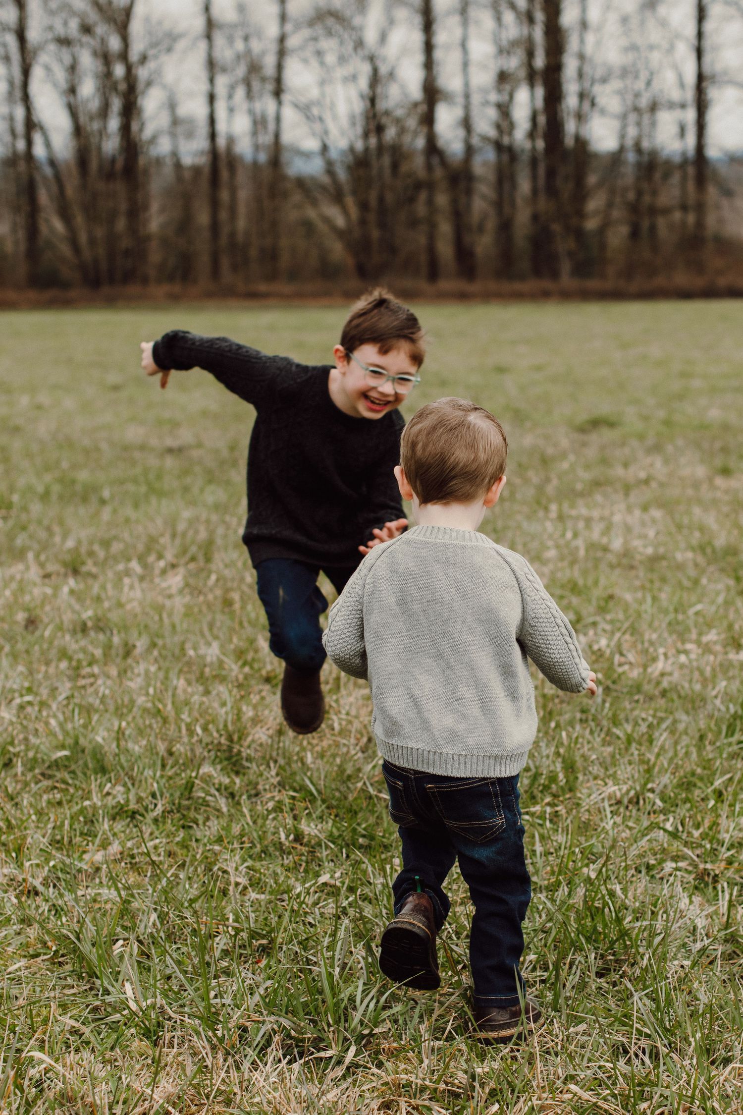 Two children playing and running together in a grassy field with bare trees in the background.