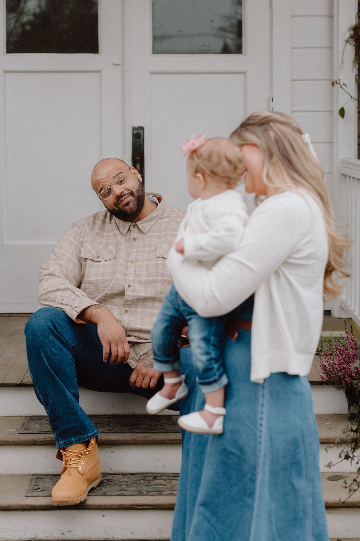 A man sits on porch steps smiling at a woman holding a toddler girl outside a white house.