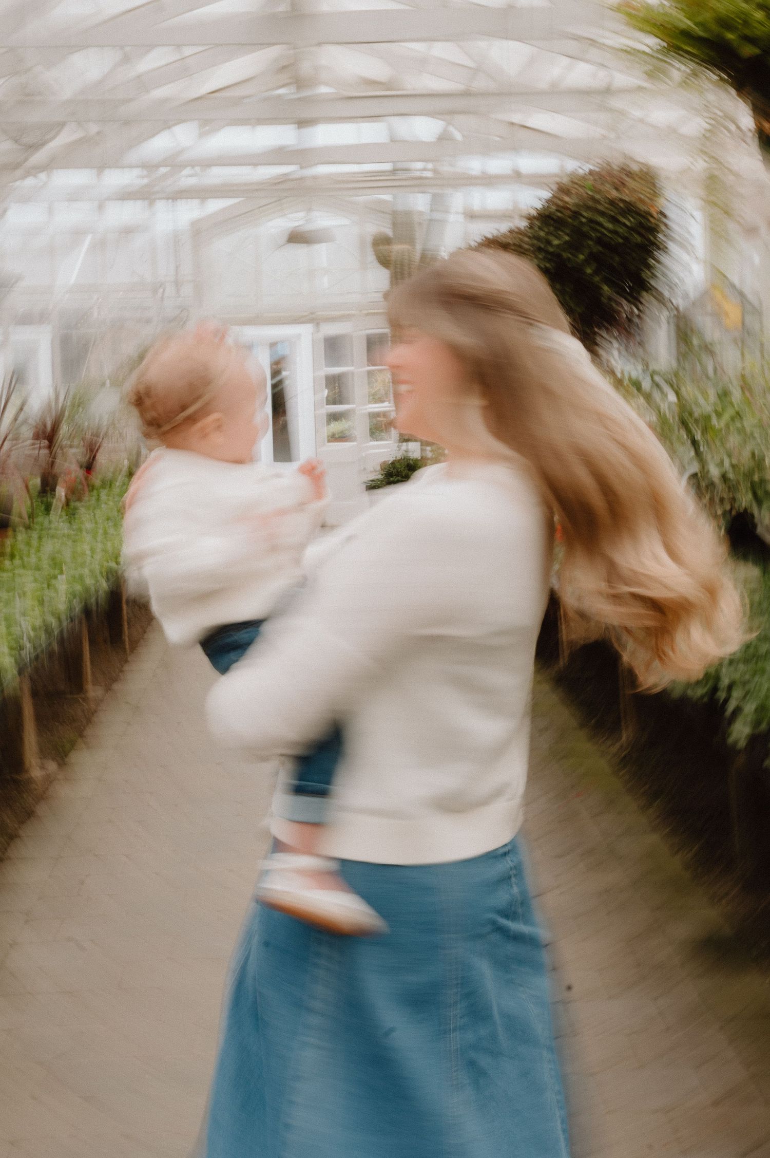 A joyful mother holds her baby in a sunlit greenhouse, her hair flowing as she smiles warmly in a blue skirt.