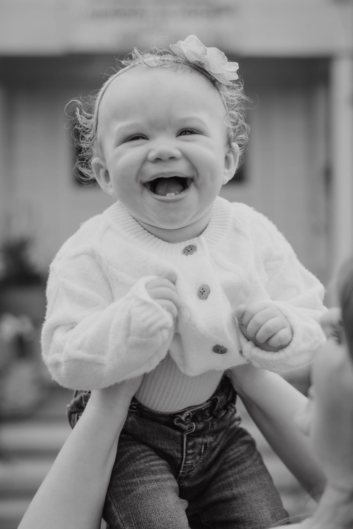 Black and white photo of a joyful laughing baby girl in white cardigan and jeans, held up by adult hands.