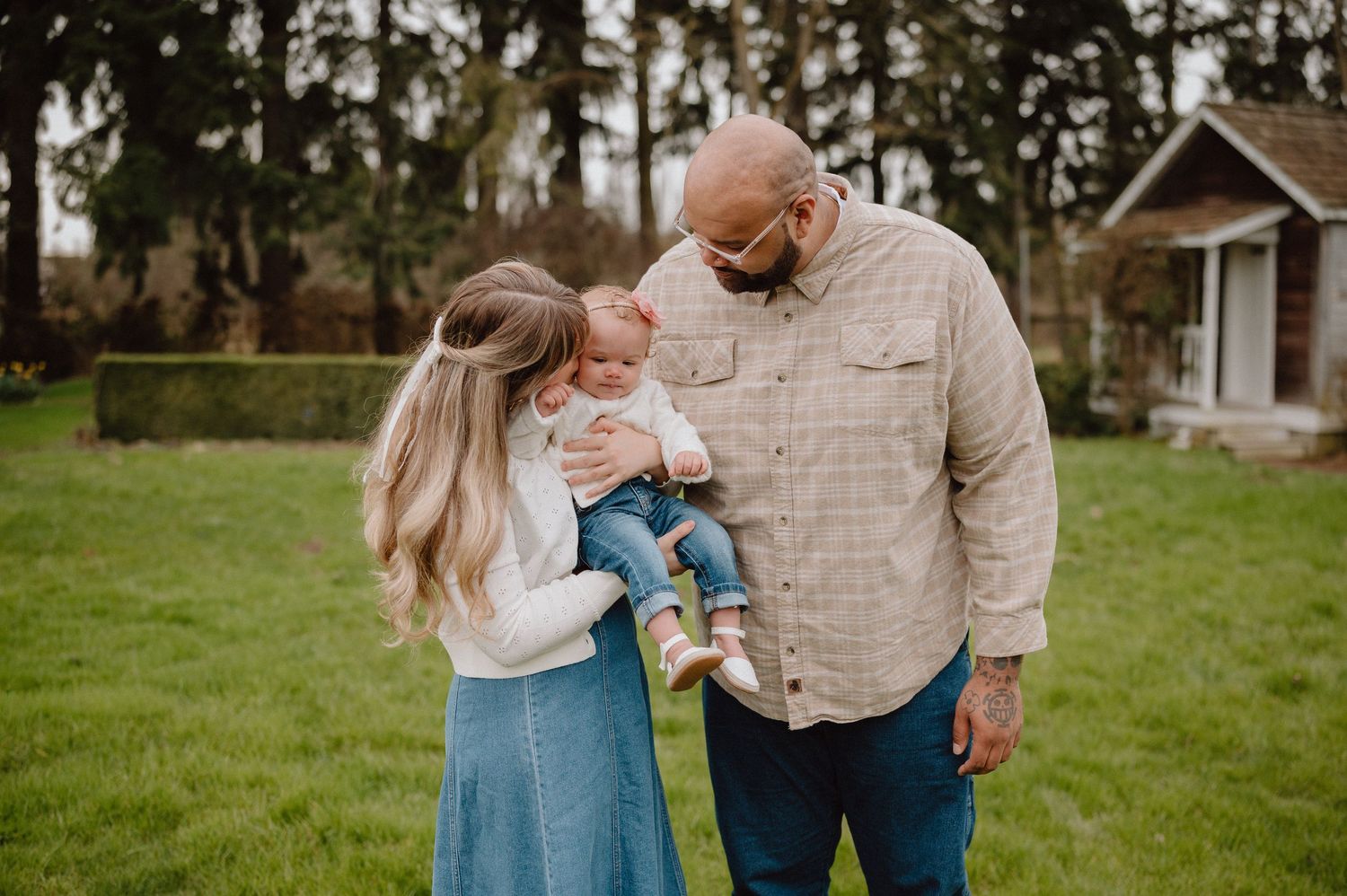 A couple shares a tender moment with their baby outdoors on a green lawn near a charming cottage.