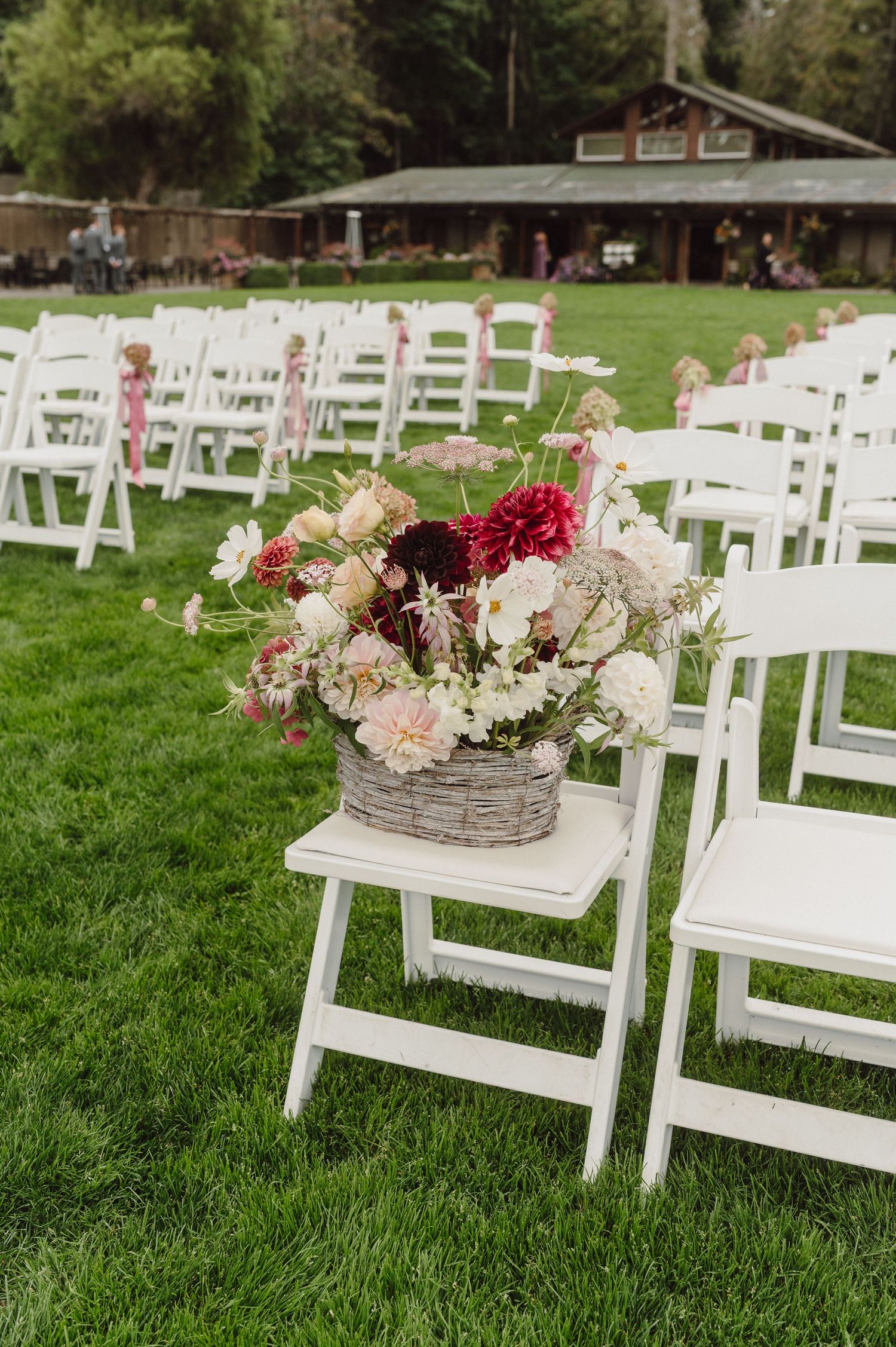 White wooden folding chairs set up on green grass for an outdoor wedding ceremony with floral arrangements.