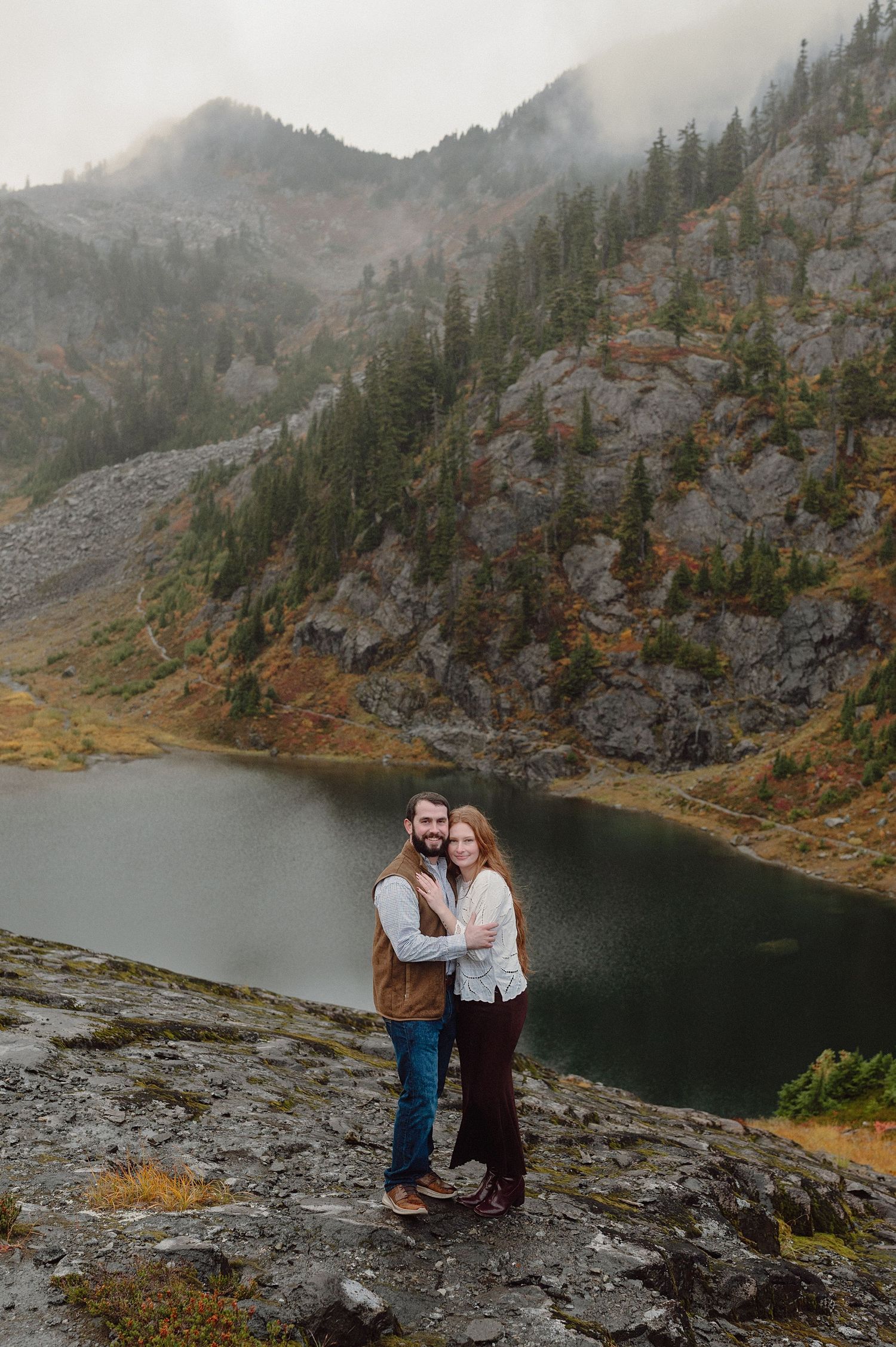 A couple stands on rocky terrain overlooking a misty mountain lake surrounded by evergreen forests and cliffs.