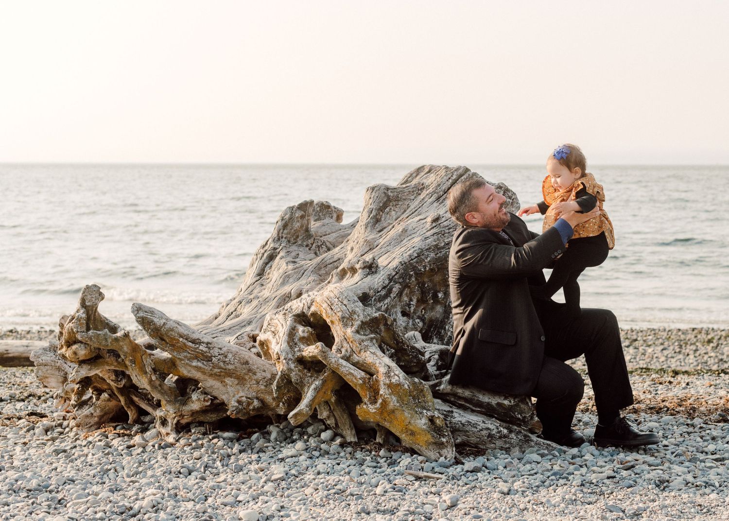 A parent and child enjoy a moment together on a rocky beach next to driftwood as waves roll in the background.