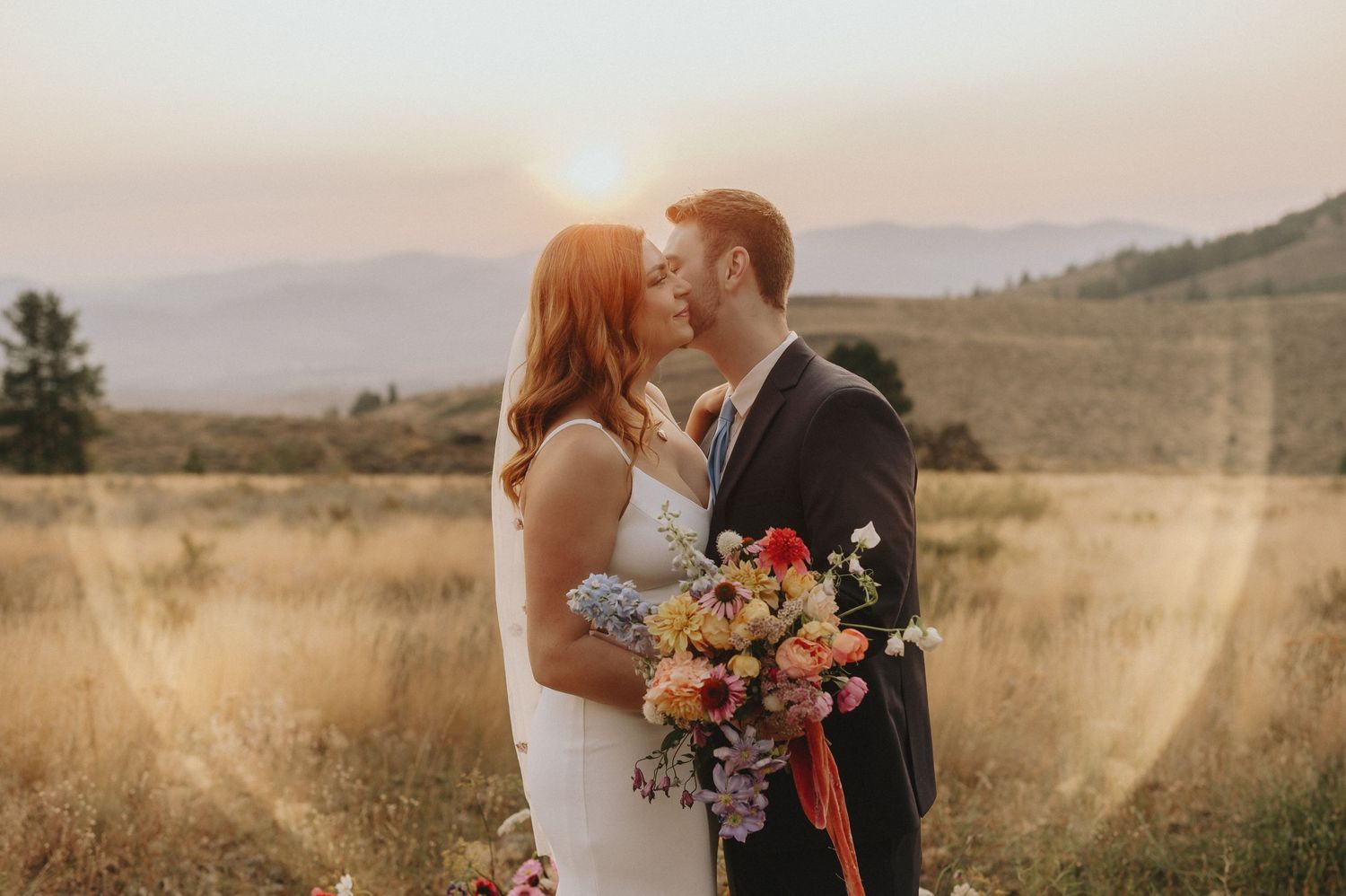 A romantic wedding couple shares a tender moment in a golden field at sunset with mountains in the background.