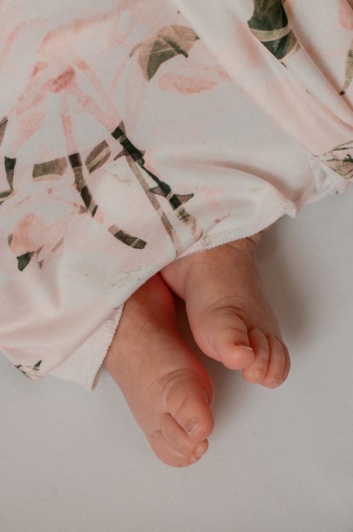 Close-up of tiny baby feet peeking out from under a pink floral dress against a white background.