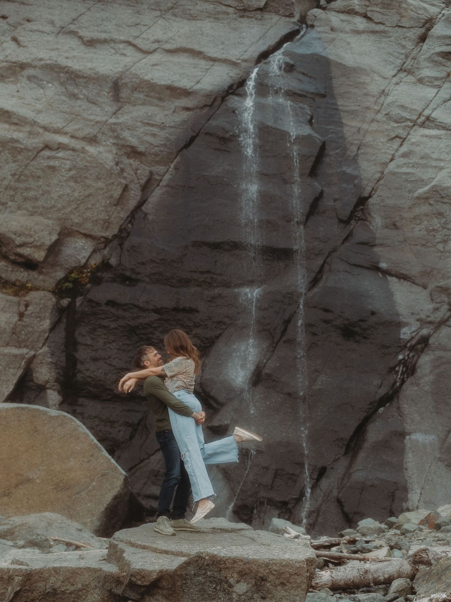 A couple embraces in front of a dramatic waterfall cascading down tall rocky cliffs.