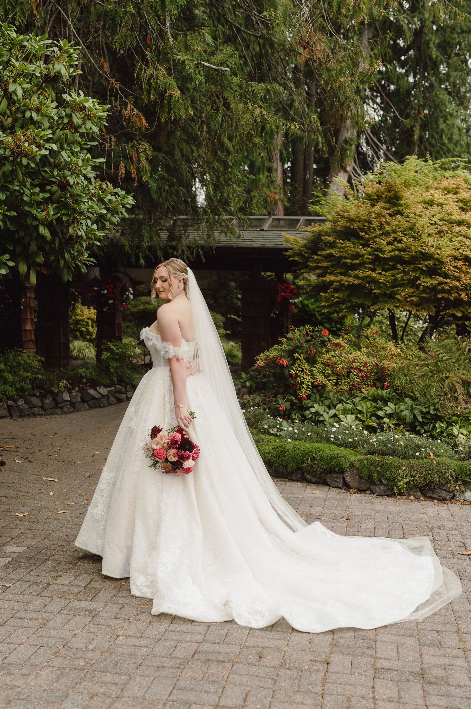 A bride in a flowing white wedding gown with a long train poses with a vibrant bouquet on a stone paved path.