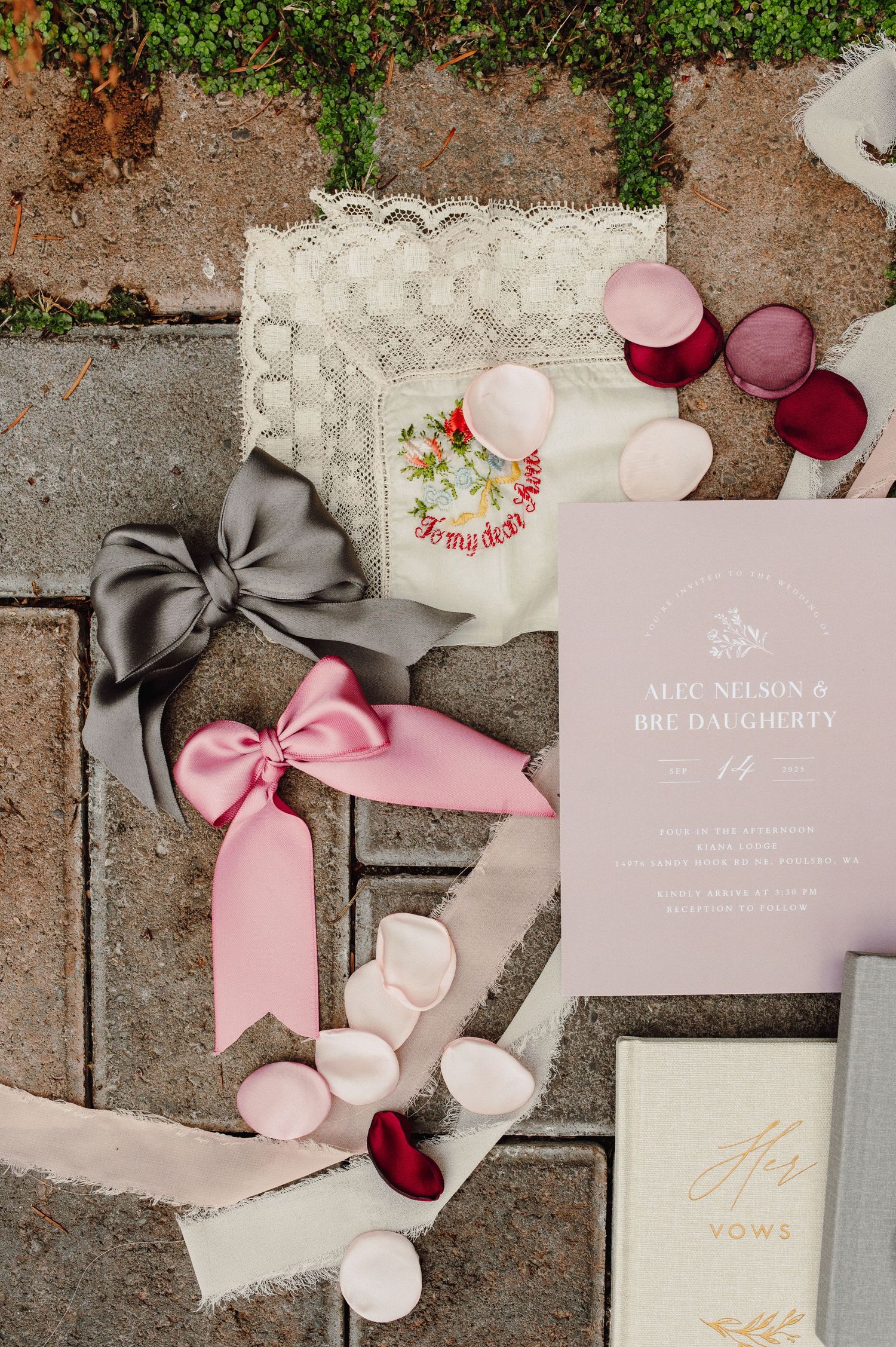 Wedding invitation suite in blush pink and gray with silk ribbons and rose petals on a textured surface.
