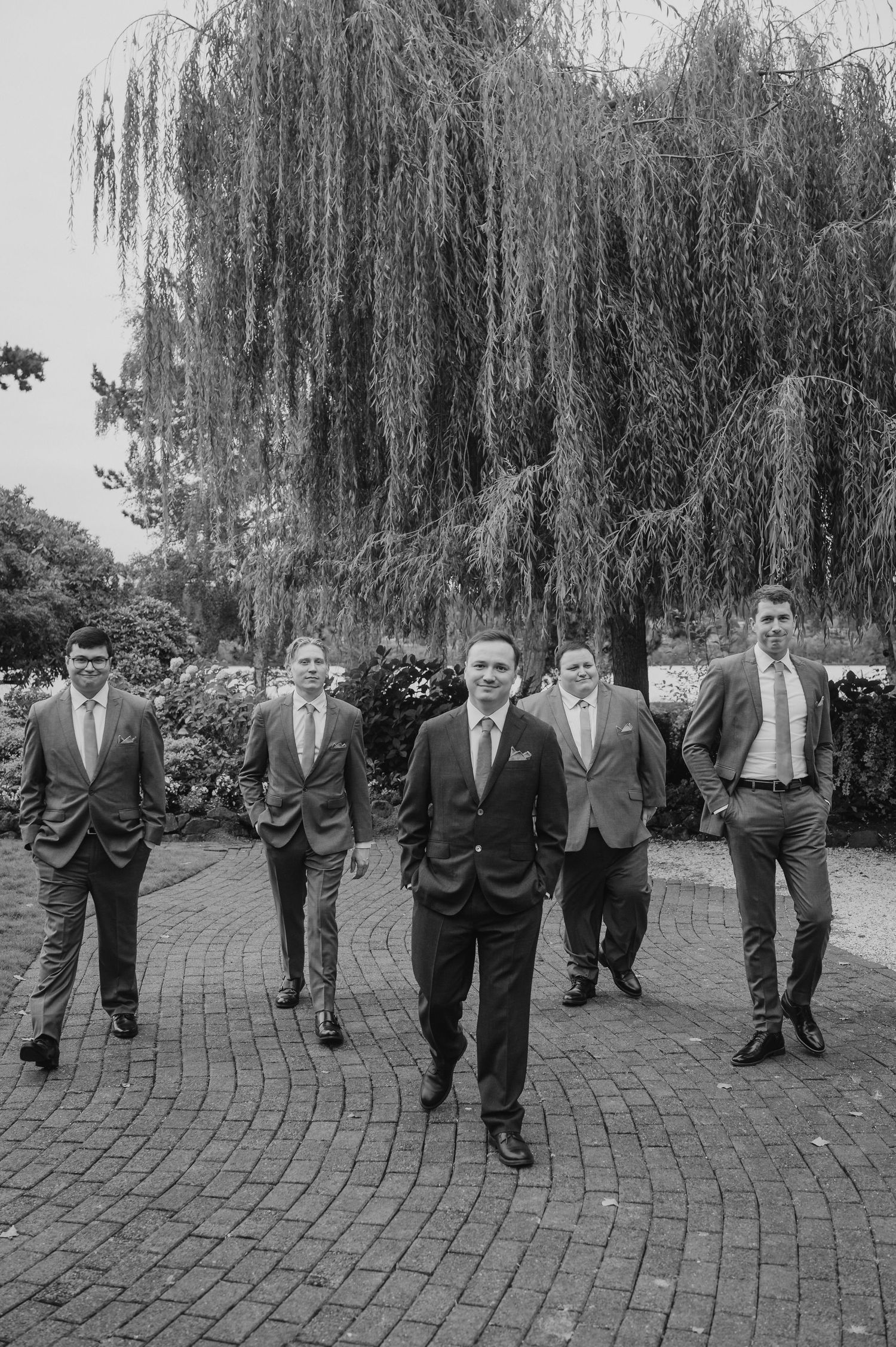 A group of men in suits walking together on a brick path with a dramatic weeping willow tree in the background.