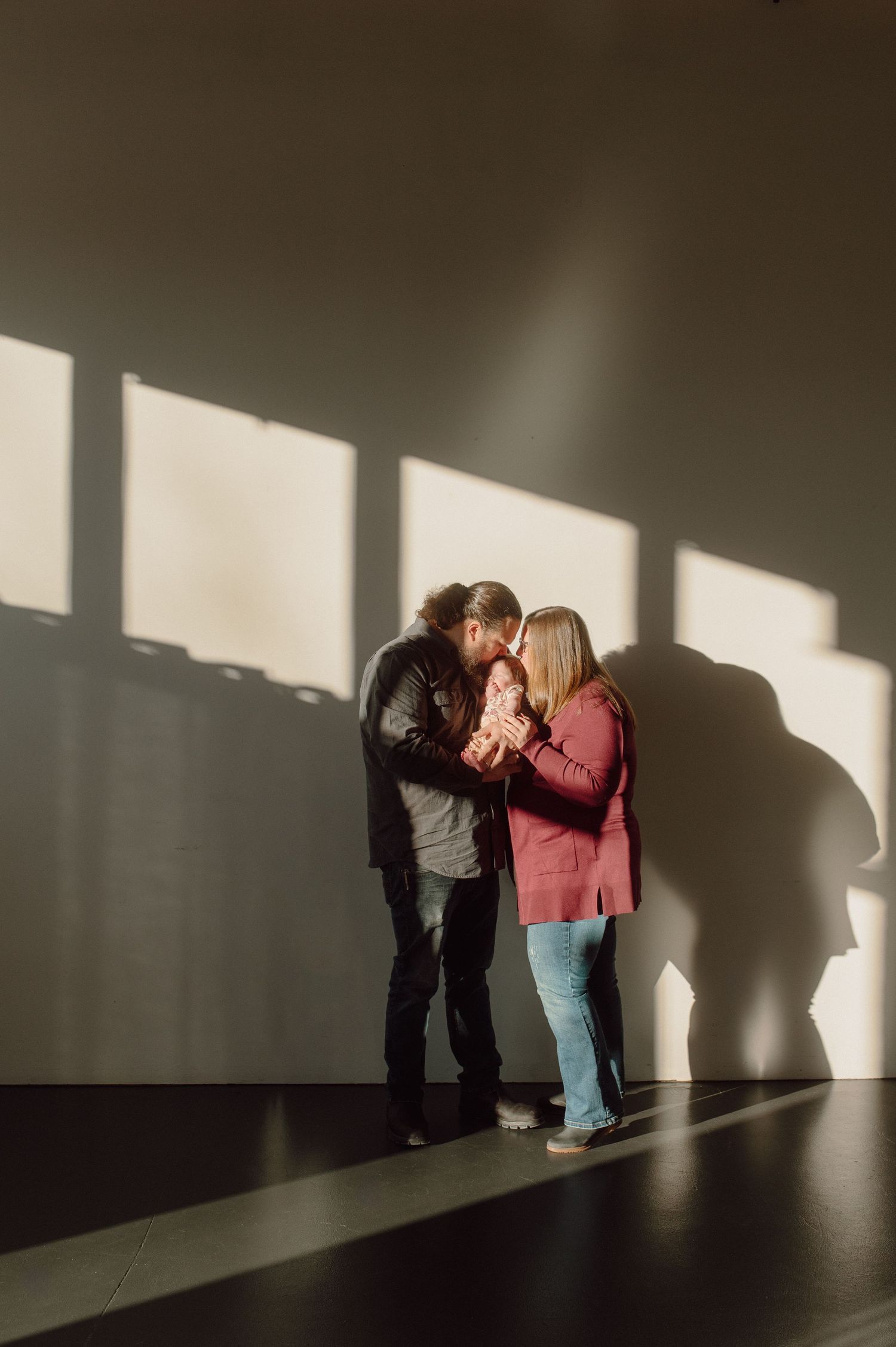 A couple stands together in a room with dramatic window light casting shadows on a wall while holding hands together.