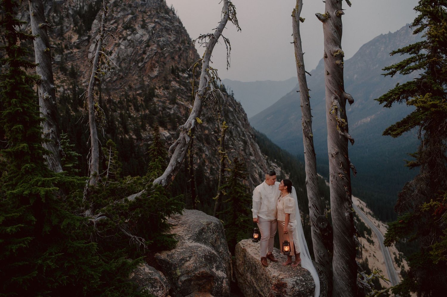 Two people stand on a rocky mountain ledge surrounded by birch trees and misty mountain views at sunset.