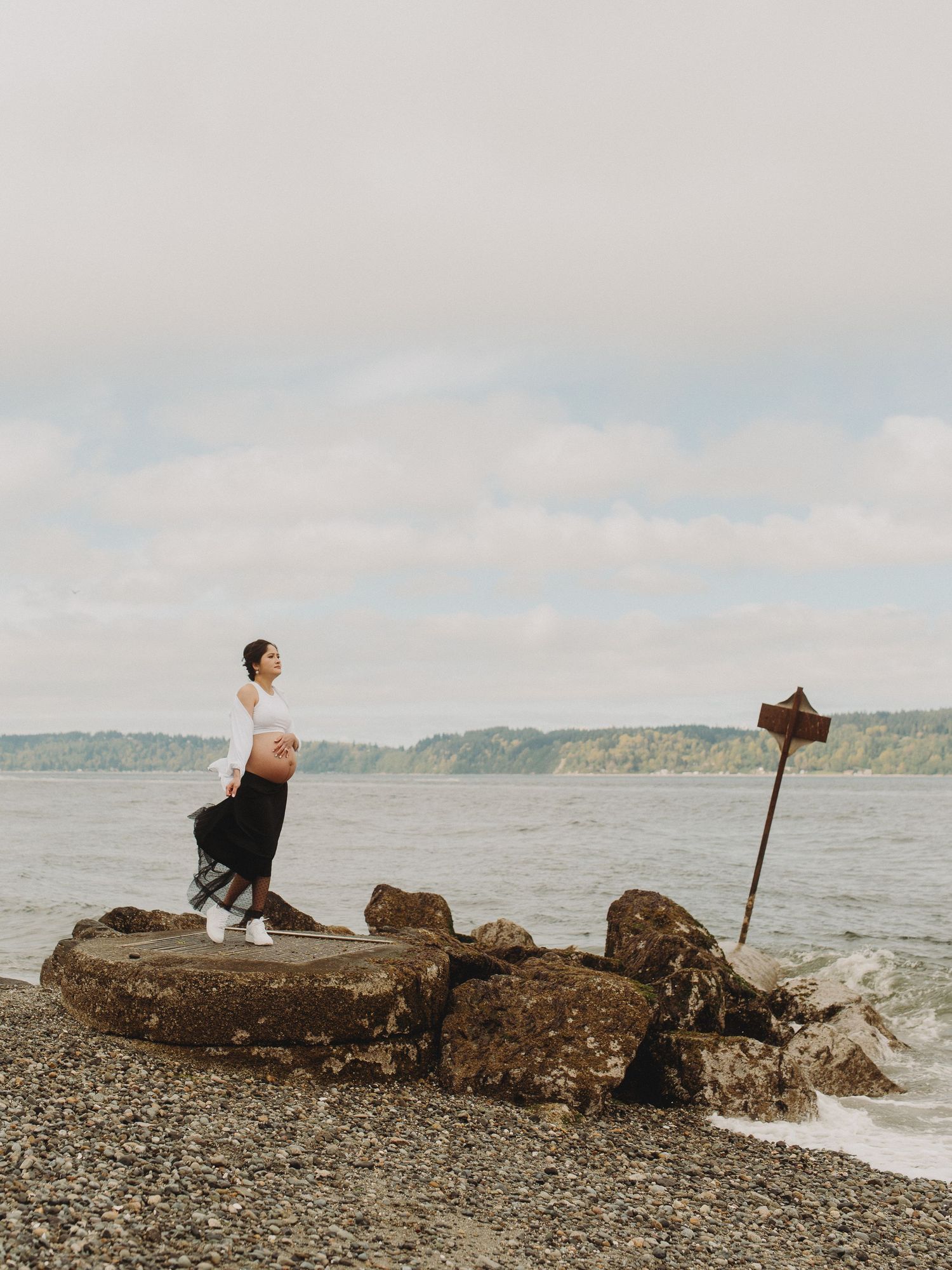 Person standing on rocky beach outcrop overlooking water with cloudy sky.