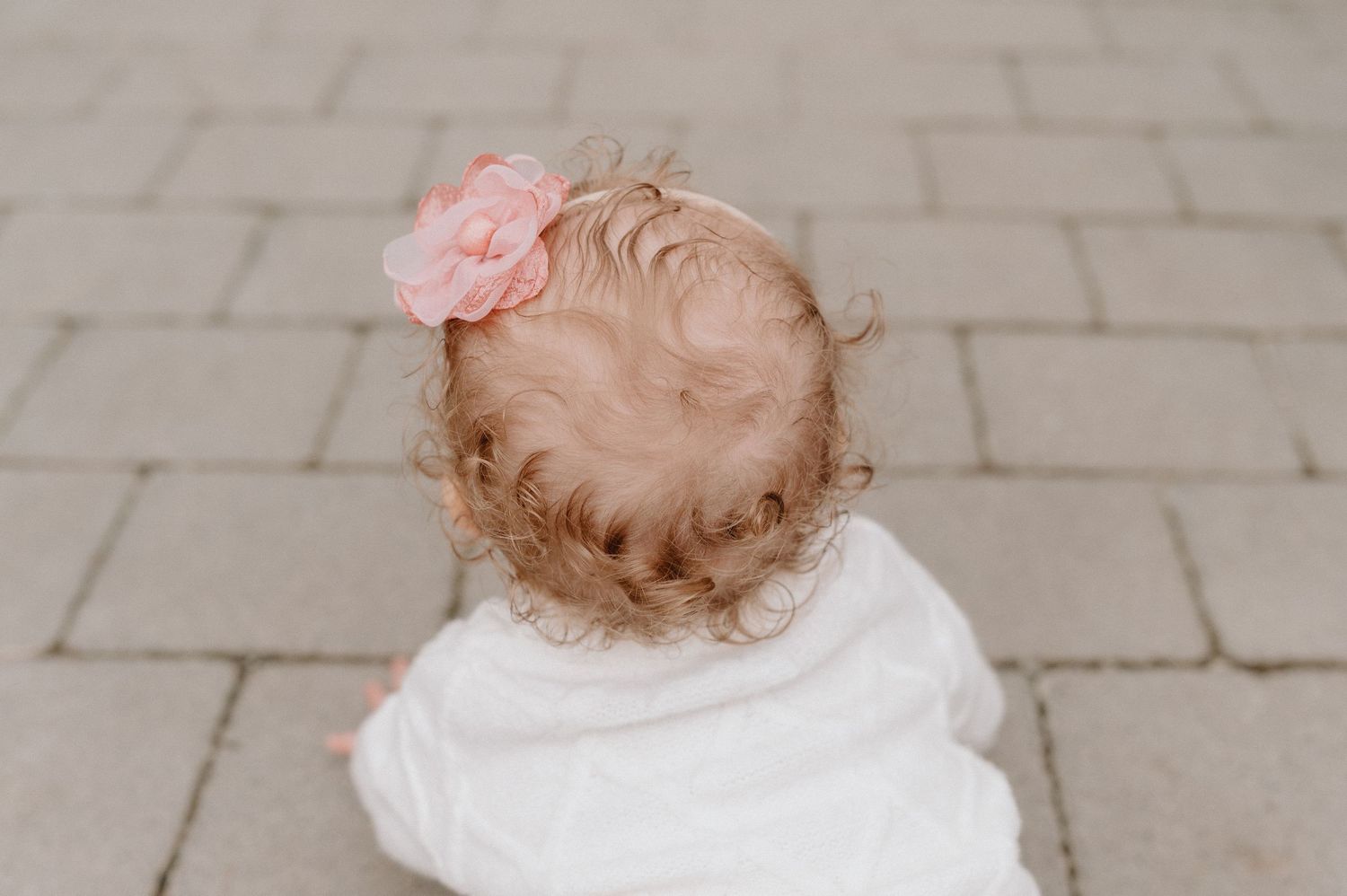 Overhead view of a baby girl with curly hair and a pink flower clip, sitting on a stone paved path.