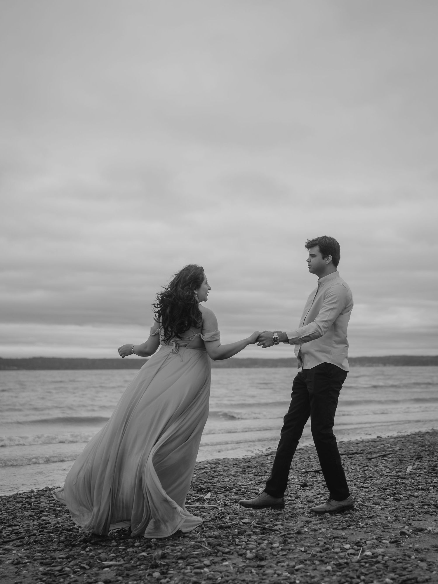 A couple dances romantically on a rocky beach, the woman in a flowing gown, in a dreamy black and white photo.