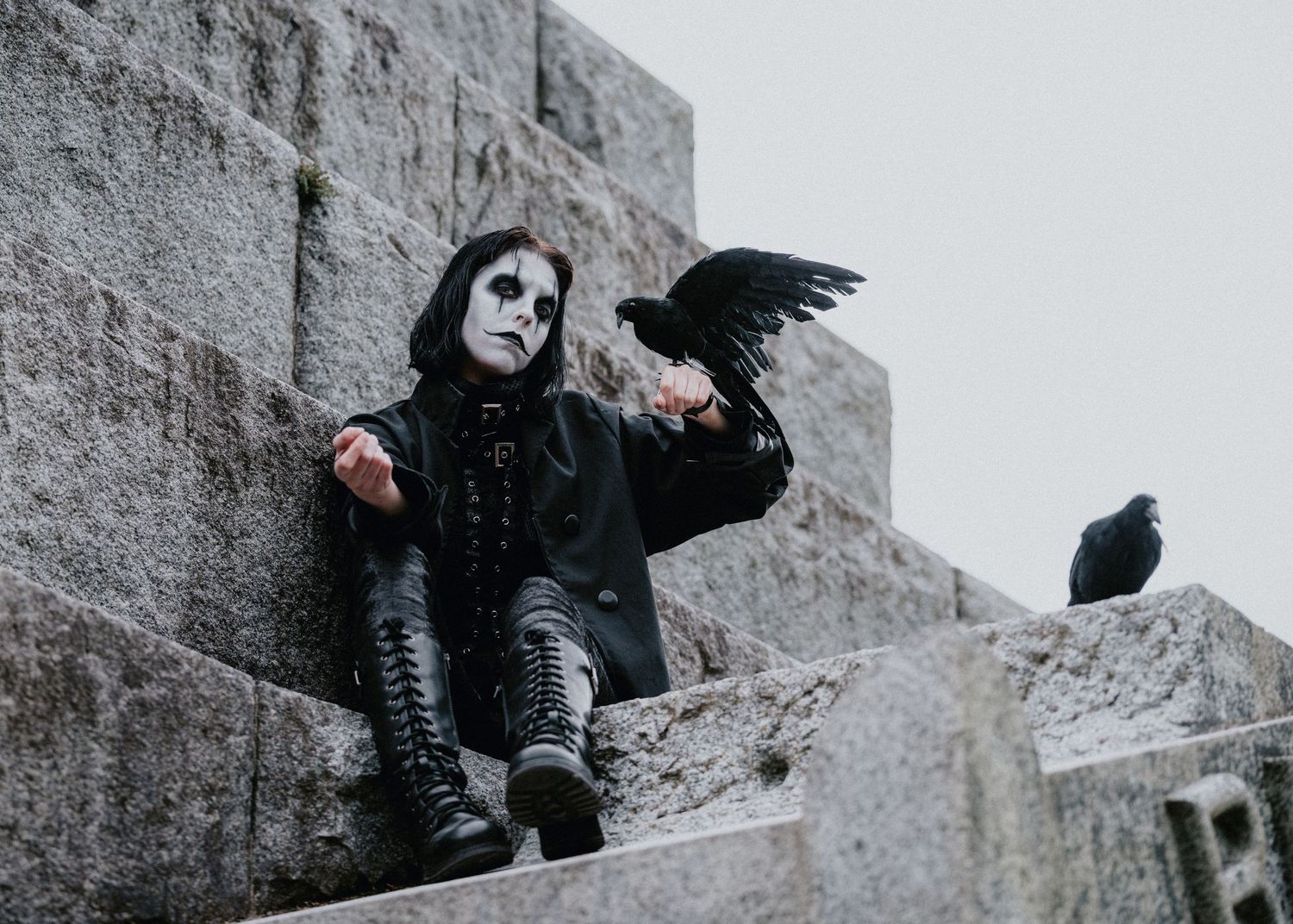 A dramatic black and white photo series showing a figure in dark clothing posing with a crow on stone steps.