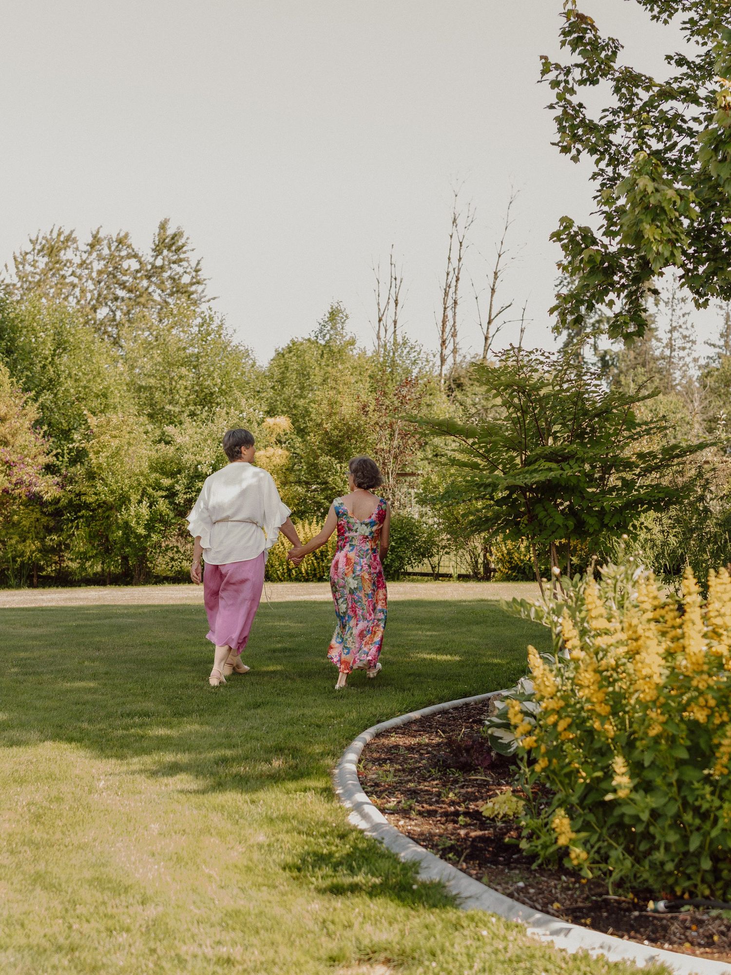 Two people running together across a sunny lawn bordered by yellow flowers and trees.