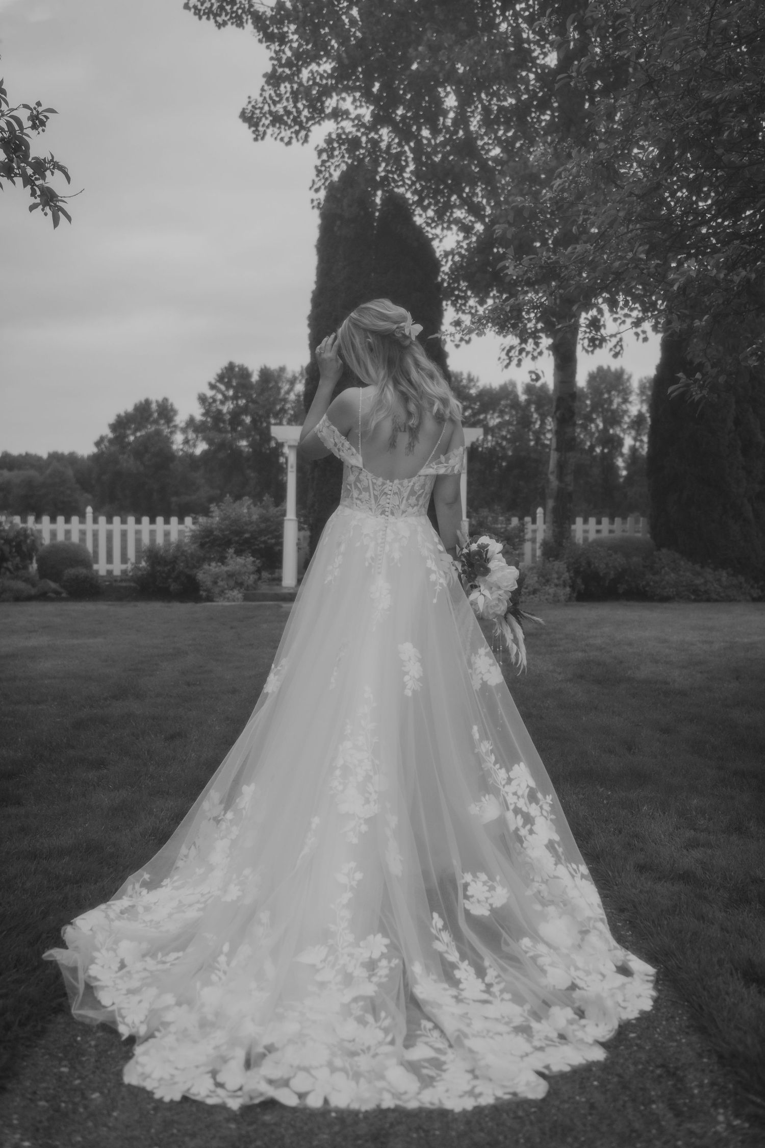 Elegant bridal gown with floral lace details photographed from behind in a garden setting with white picket fence.