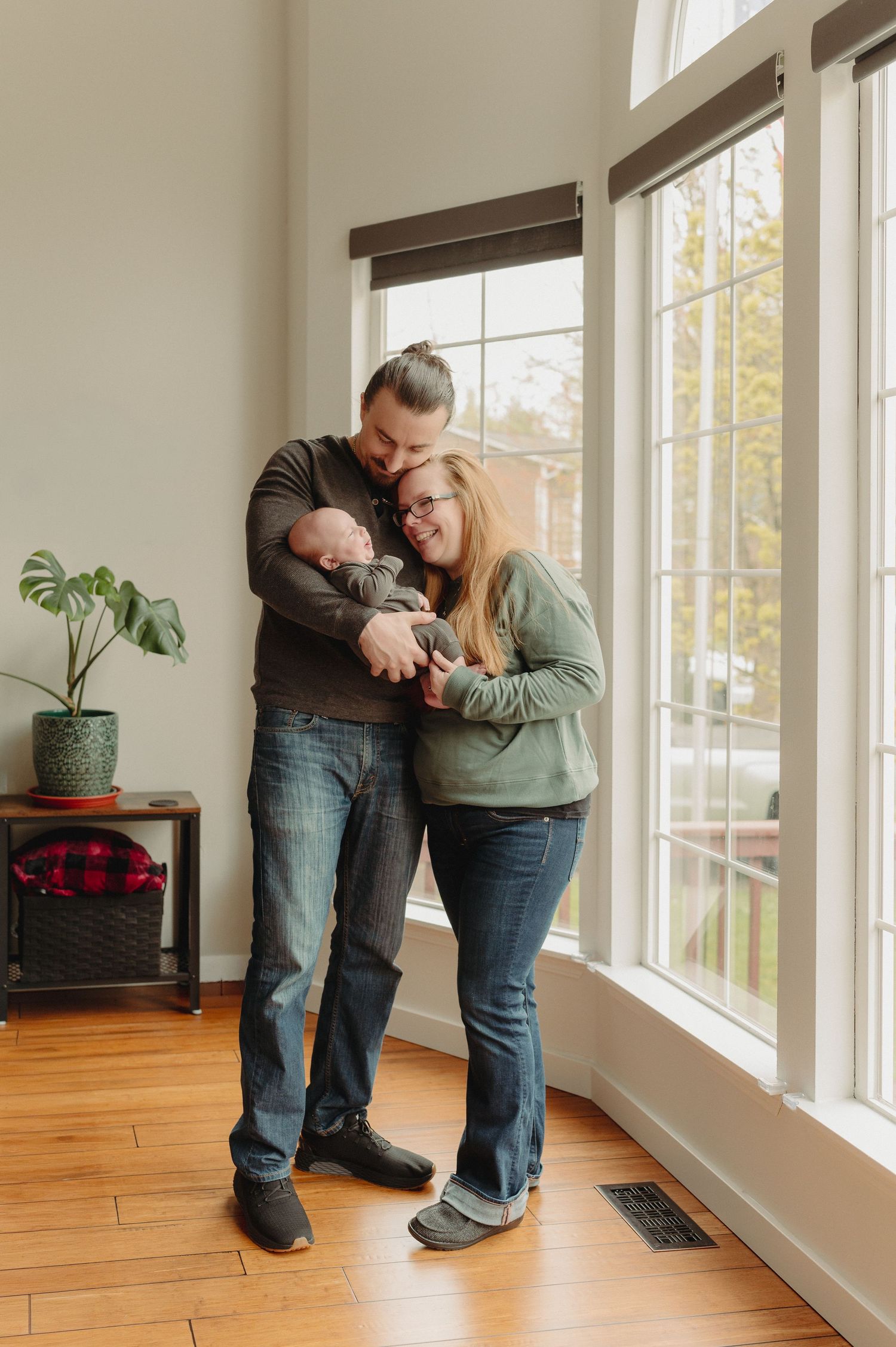 A couple stands by large windows holding their newborn baby, smiling warmly in a bright, modern living room.