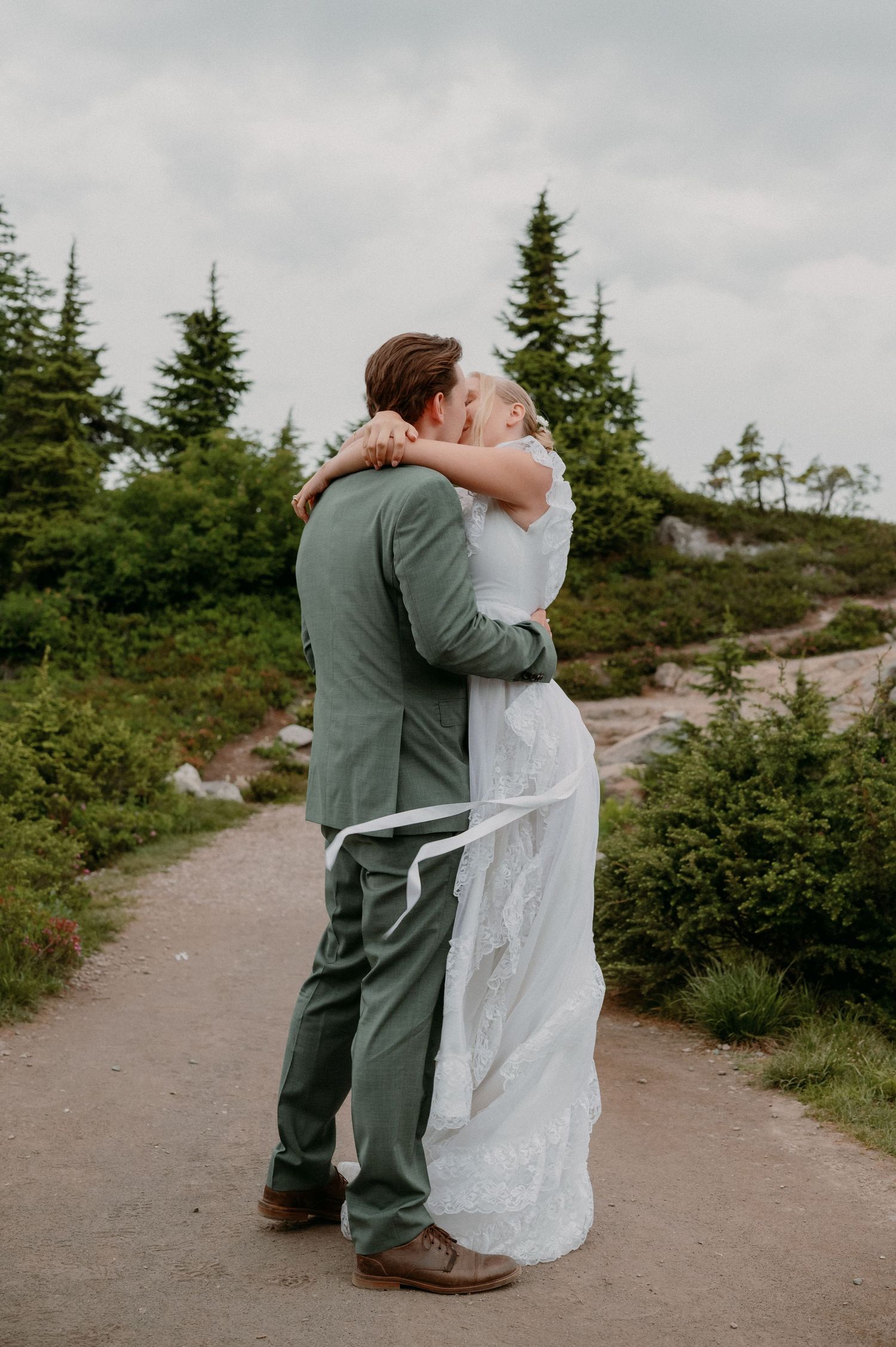 Bride and groom share a romantic kiss on a scenic mountain path surrounded by evergreen trees on their wedding day.