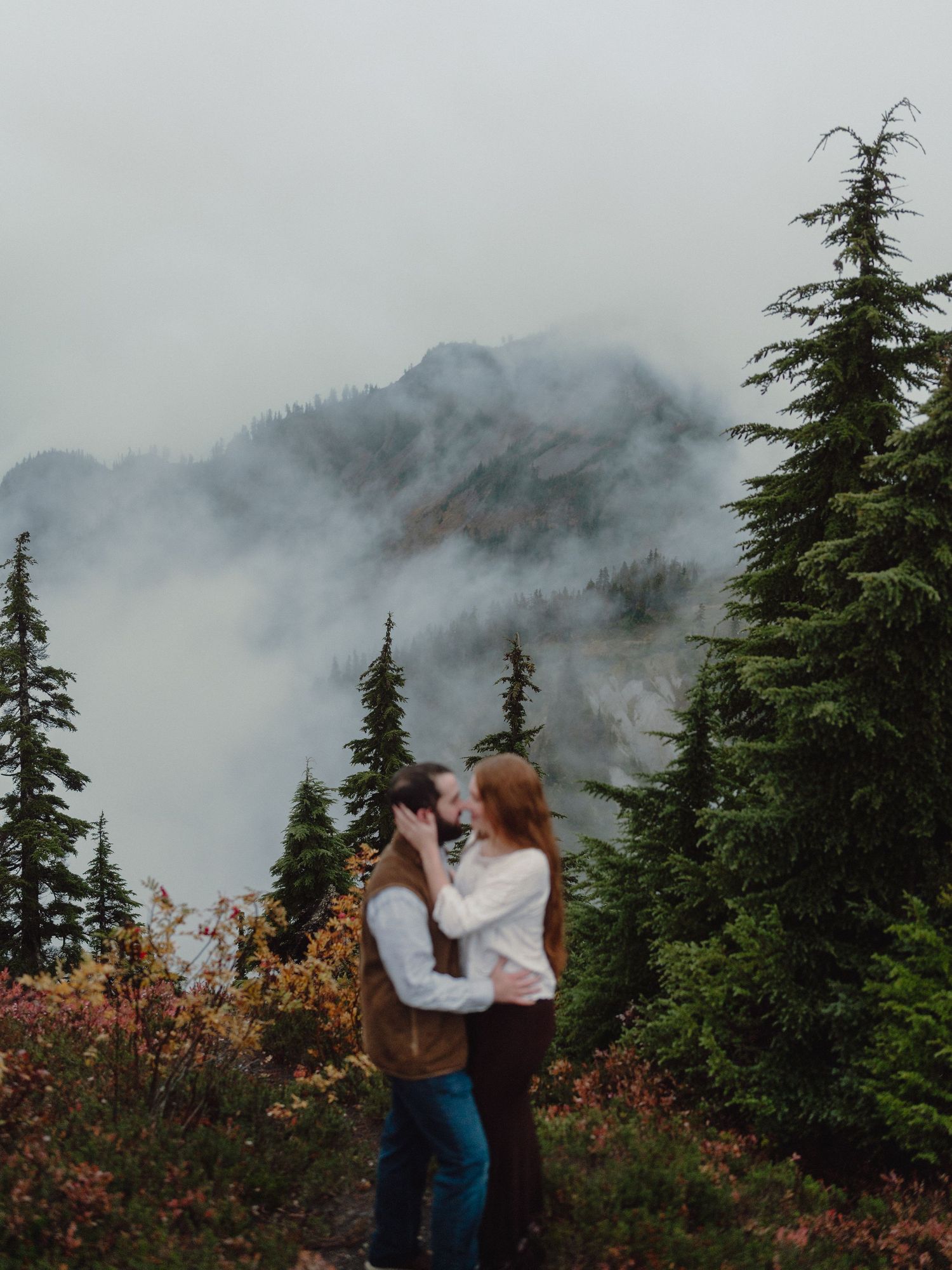 Couple embraces while taking photos in misty mountain landscape with evergreen trees and fall foliage.