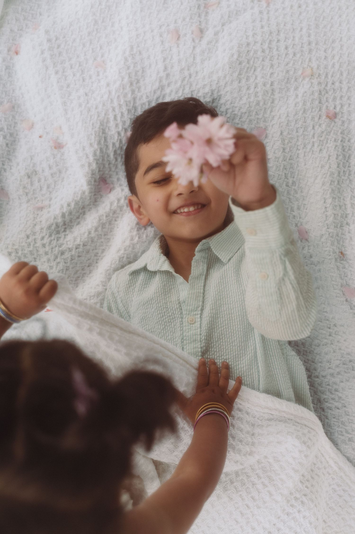 Child lying on white blanket holds pink flower while smiling playfully.