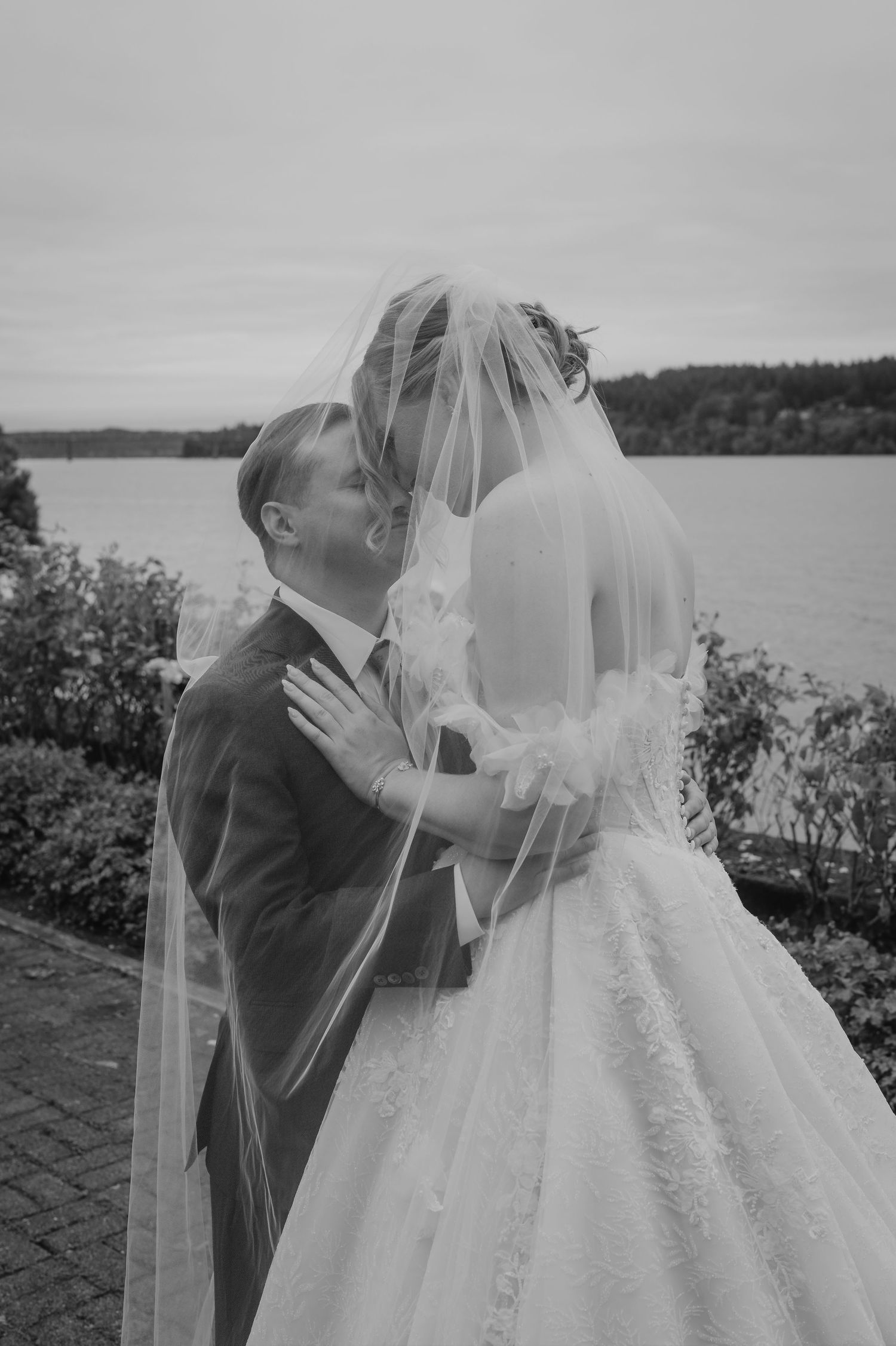 A romantic black and white wedding moment captured by a lake with trees in the background.