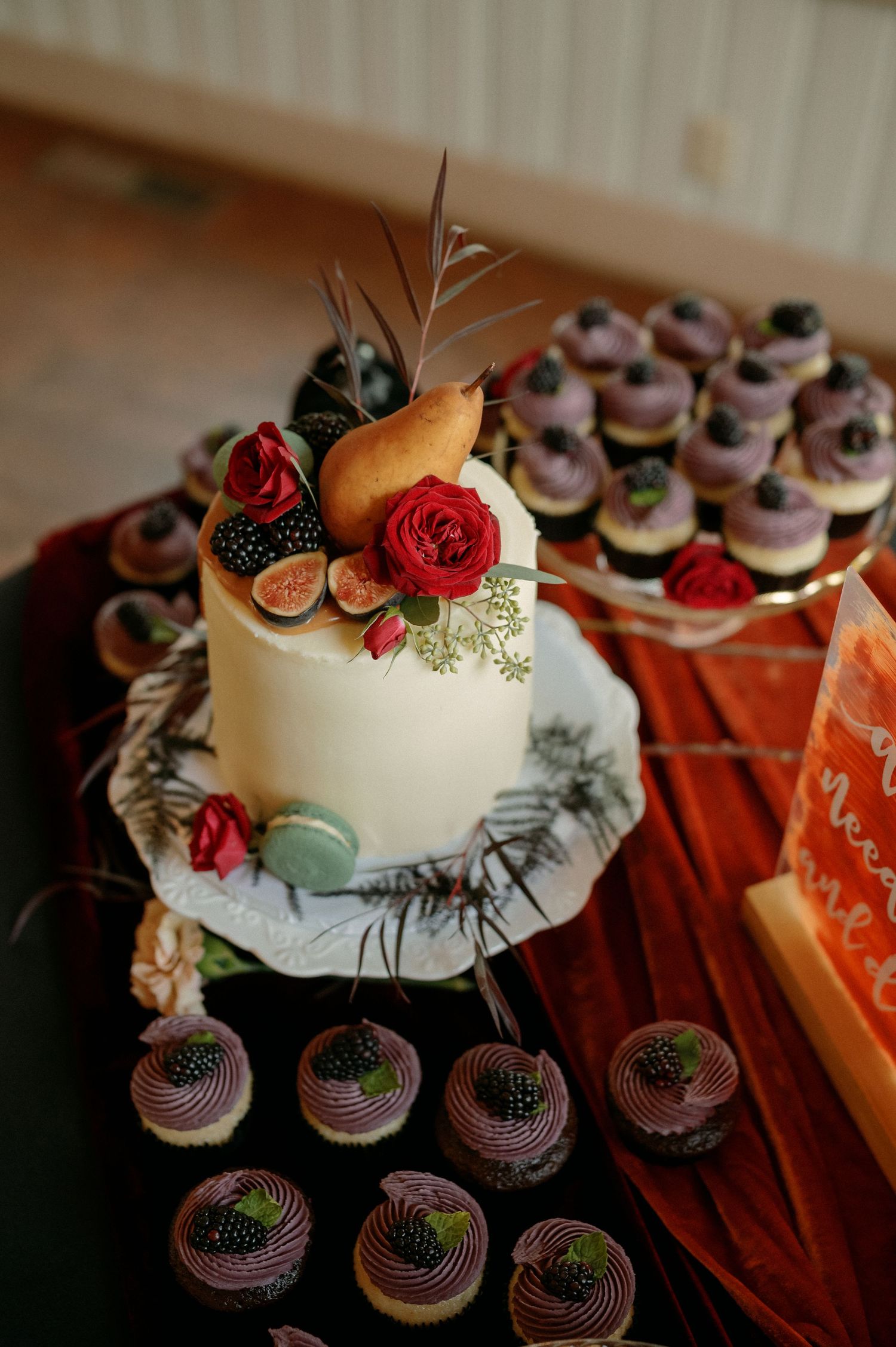 White wedding cake decorated with fresh fruits, roses and figs displayed alongside purple frosted cupcakes on a wooden table.