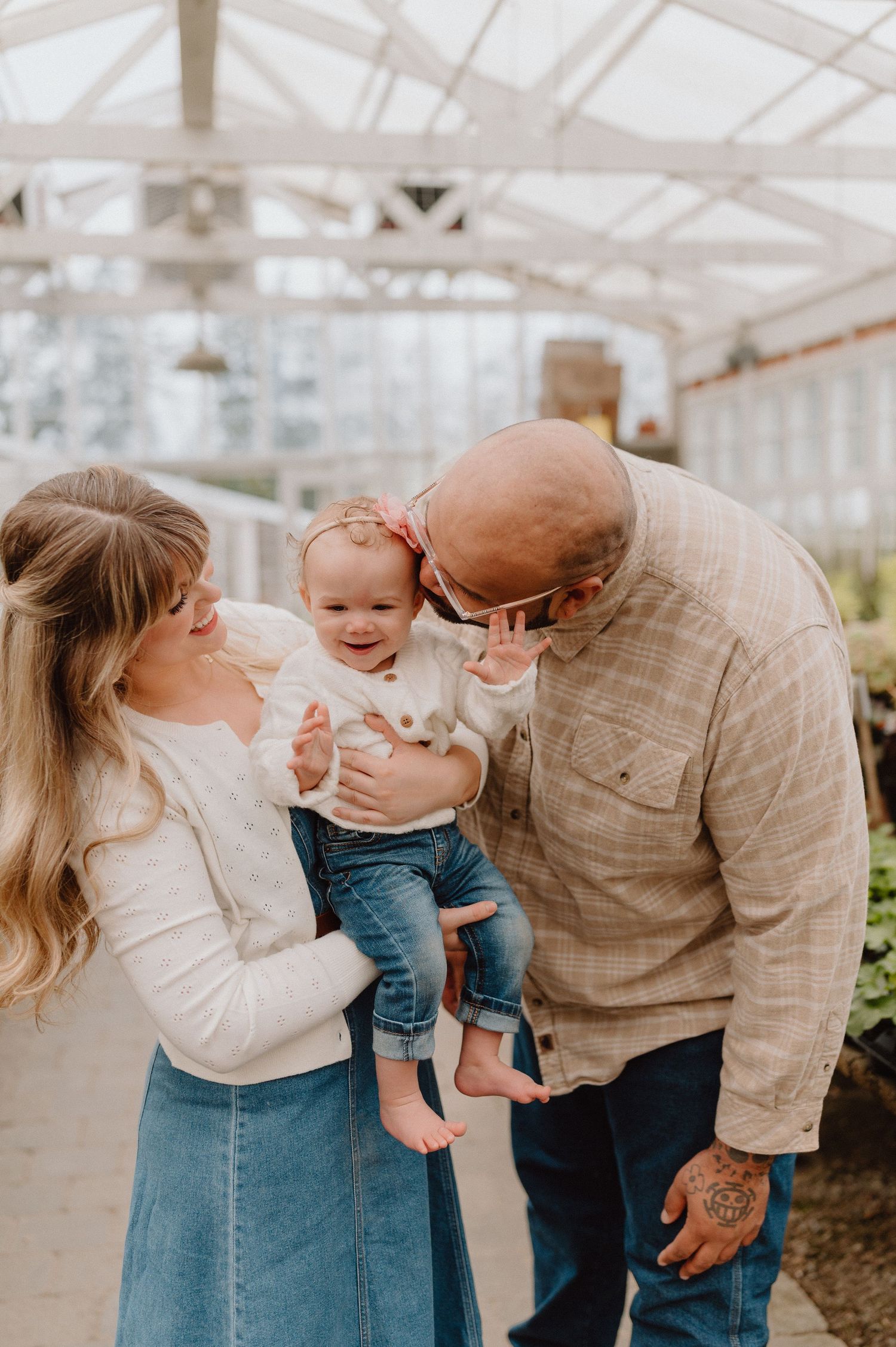 A couple smiles at their happy baby girl in a bright greenhouse, creating a warm family moment.