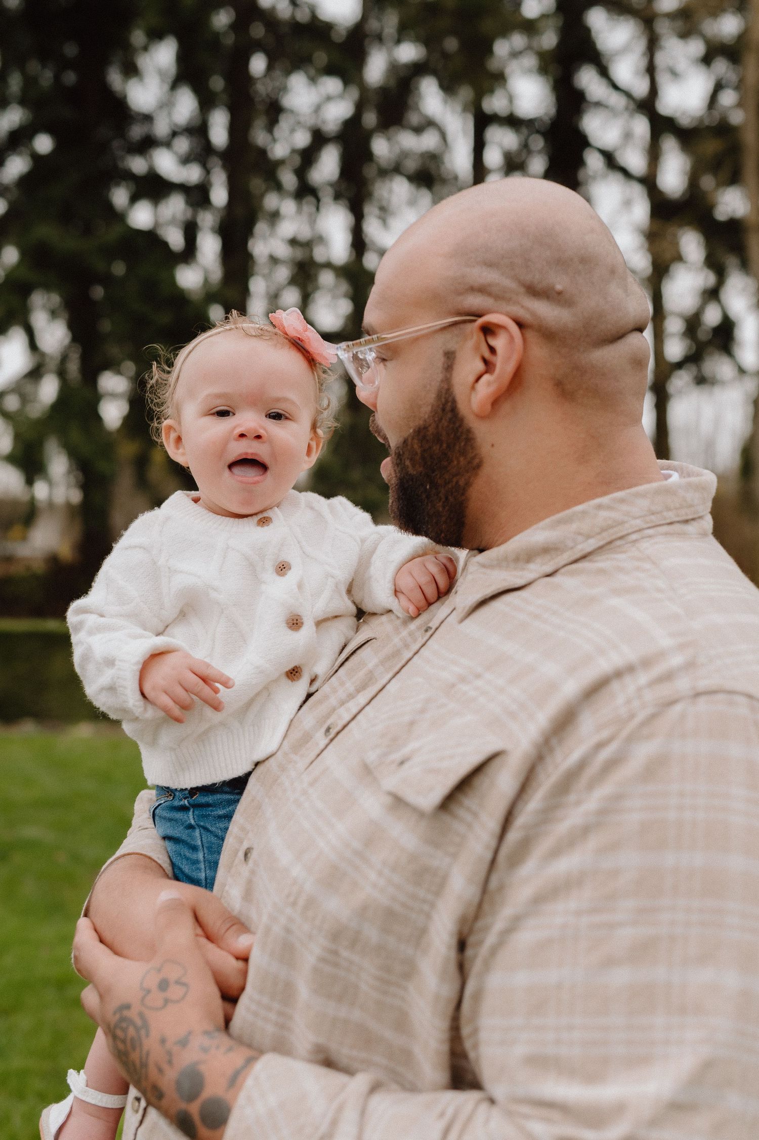 Bald bearded father with glasses holds laughing baby girl in white sweater outdoors near green trees.