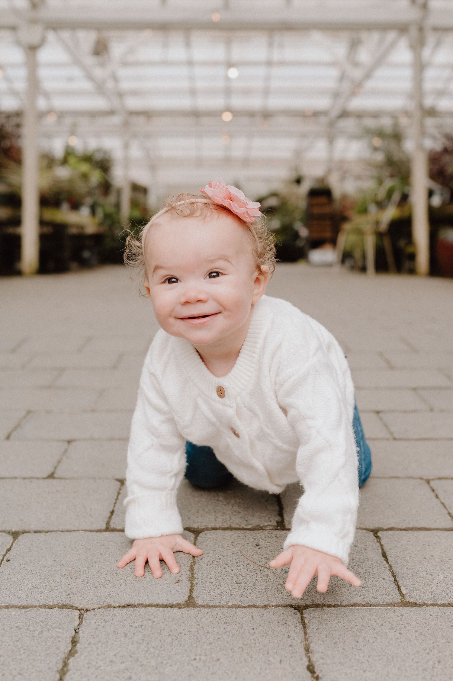 An adorable baby girl in a white knit sweater and pink bow crawls on a stone path inside a greenhouse.