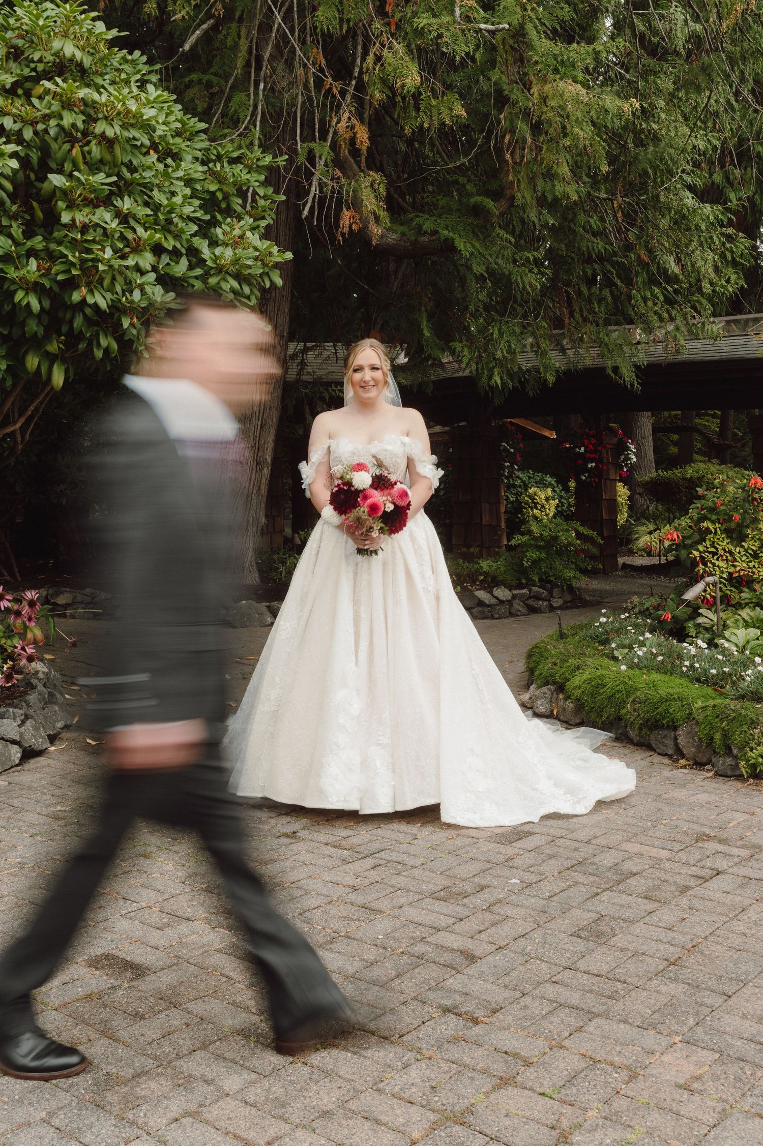 A wedding scene showing motion-blurred figures walking past a person in a white wedding gown on a cobblestone path.