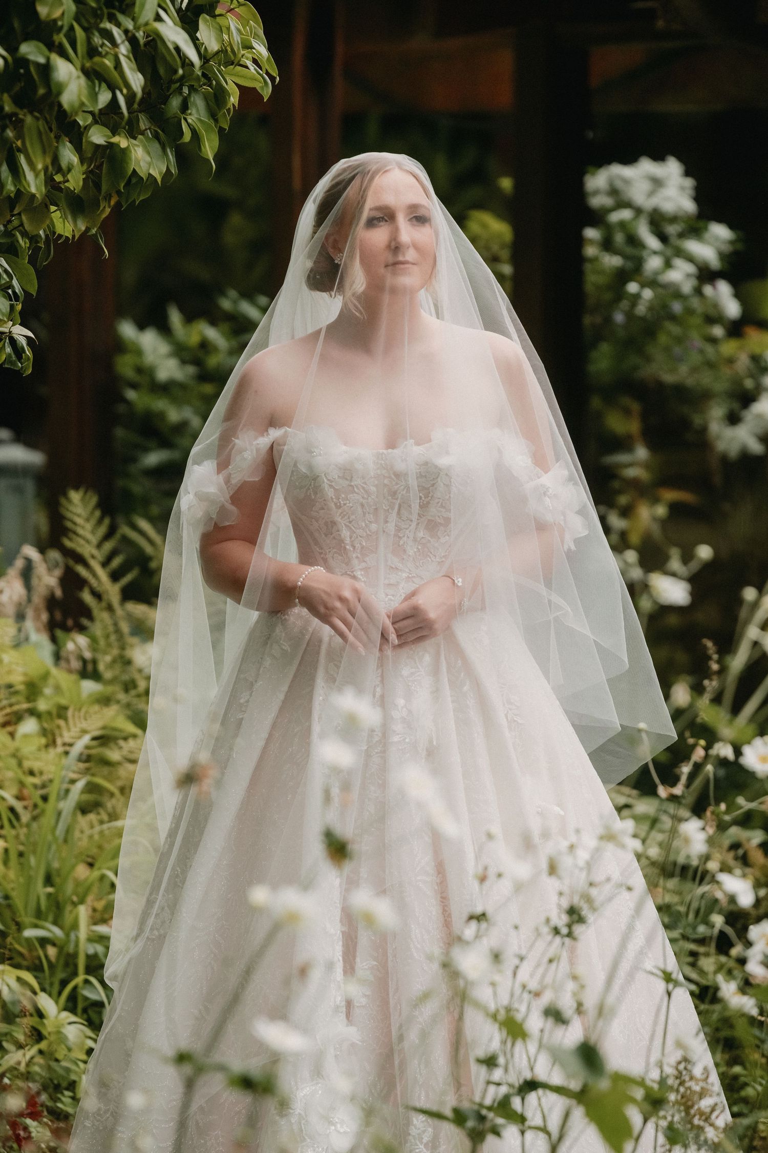 A bride in a white lace wedding dress stands among garden wildflowers with a flowing sheer veil.